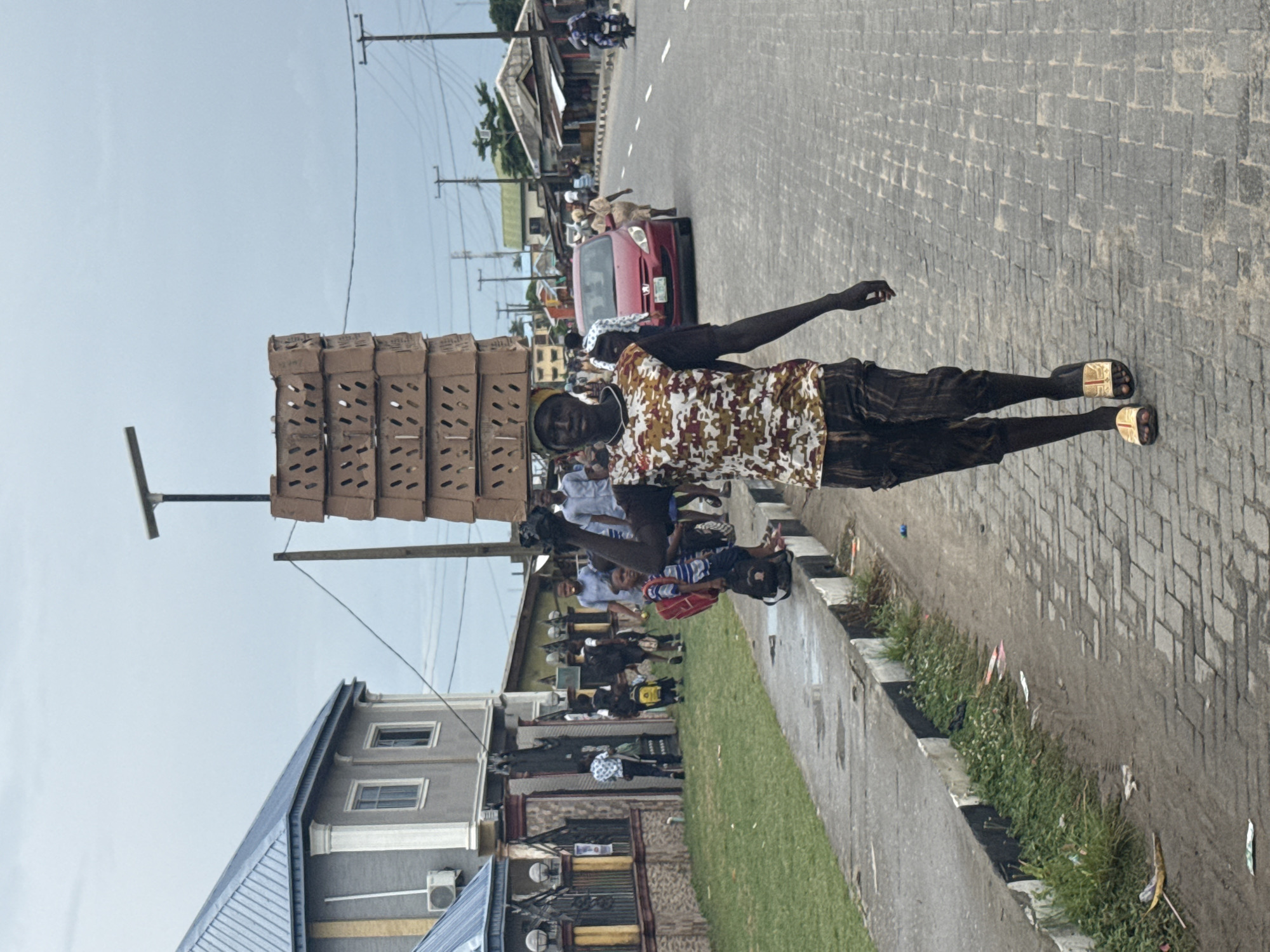 A man carries a tall stack of cardboard boxes on his head while walking along a brick road in what appears to be Badagry, Nigeria. He wears a patterned shirt and dark pants, and sandals. A crowd of people stand near a sidewalk with grass and a building to the side, and a red car is visible on the road. Overhead power lines stretch across the sky. The sky is overcast.