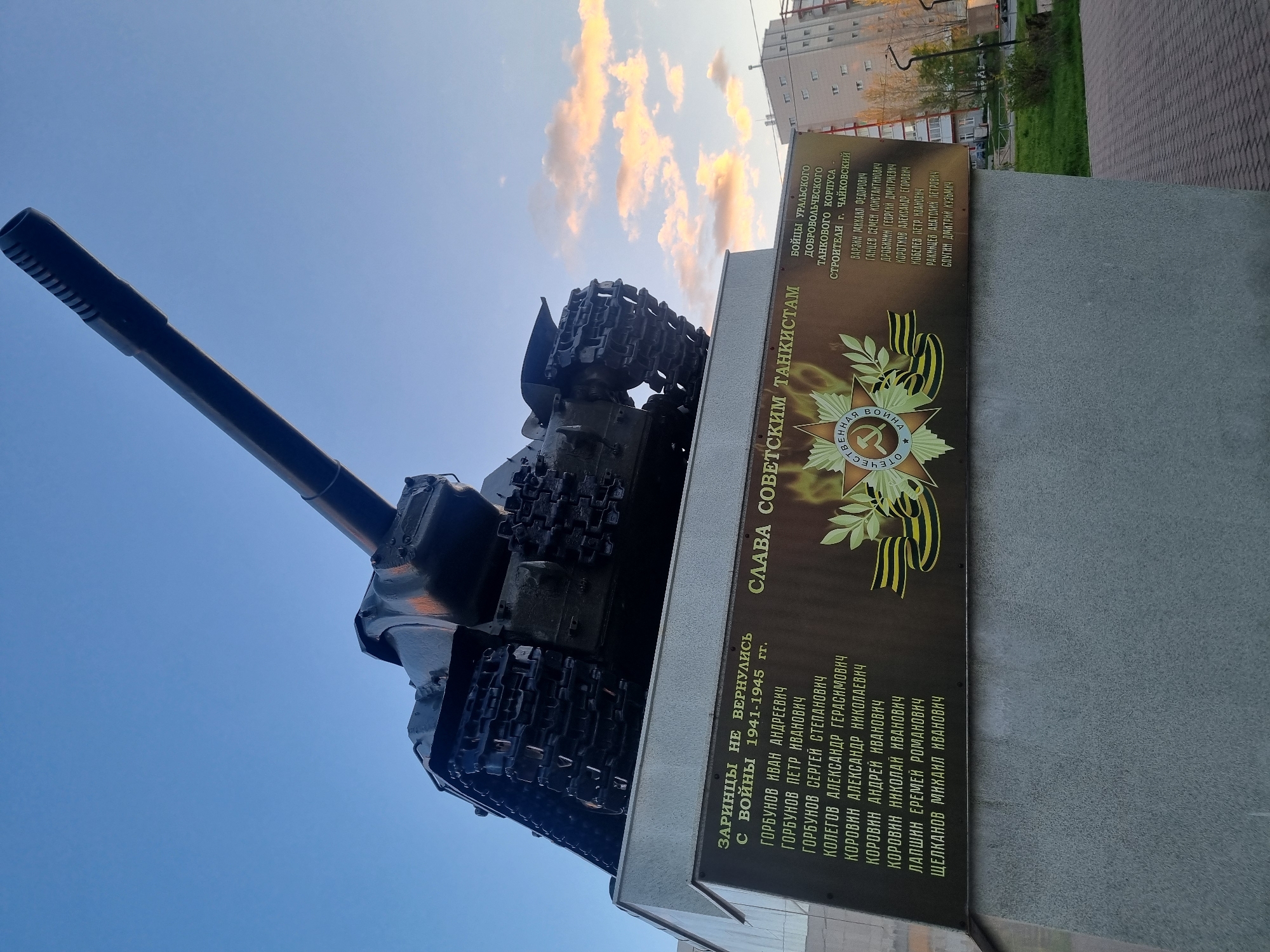 Here's a detailed description of the image:
**General Description:**
The image captures a monument dedicated to Soviet tankists in the city of Chaikovsky, Russia. The monument features a T-34 tank mounted on a concrete base. A plaque is affixed to the base, bearing inscriptions in Russian. The photo is taken from a low angle, emphasizing the tank's size and imposing presence.
**Subjects and Objects:**
* **T-34 Tank:** A Soviet medium tank of WWII. It is mounted as a central element of the memorial.
* **Concrete Base:** The tank sits upon a concrete plinth.
* **Plaque:** A rectangular dark plaque attached to the concrete base.
* **Inscription:** Text in Russian listing names (presumably those who did not return from the war) and dedications to the tankists.
* **Background:** In the background, there are buildings, trees, and the sky with scattered clouds. A portion of a paved walkway can be seen.
**Inscriptions (based on OCR):**
* "Slava Sovetskim Tankistam" (Glory to Soviet Tankists)
* "Zarintsy ne vernulis' s voyny 1941-1945 gg." (Those from Zarintsy who did not return from the war, 1941-1945)
* Lists of names, presumably local residents who served as tankists.
* "Boytsy Ural'skogo Dobrovol'cheskogo Tankovogo Korpusa, Stroiteli g. Chaykovskiy" (Fighters of the Ural Volunteer Tank Corps, Builders of Chaikovsky).
**Environment:**
* **Location:** Chaikovsky, Russia, as indicated by the inscription.
* **Time of Day:** Appears to be late afternoon or early evening, based on the soft lighting and sky colors.
* **Weather:** Clear sky with some clouds.