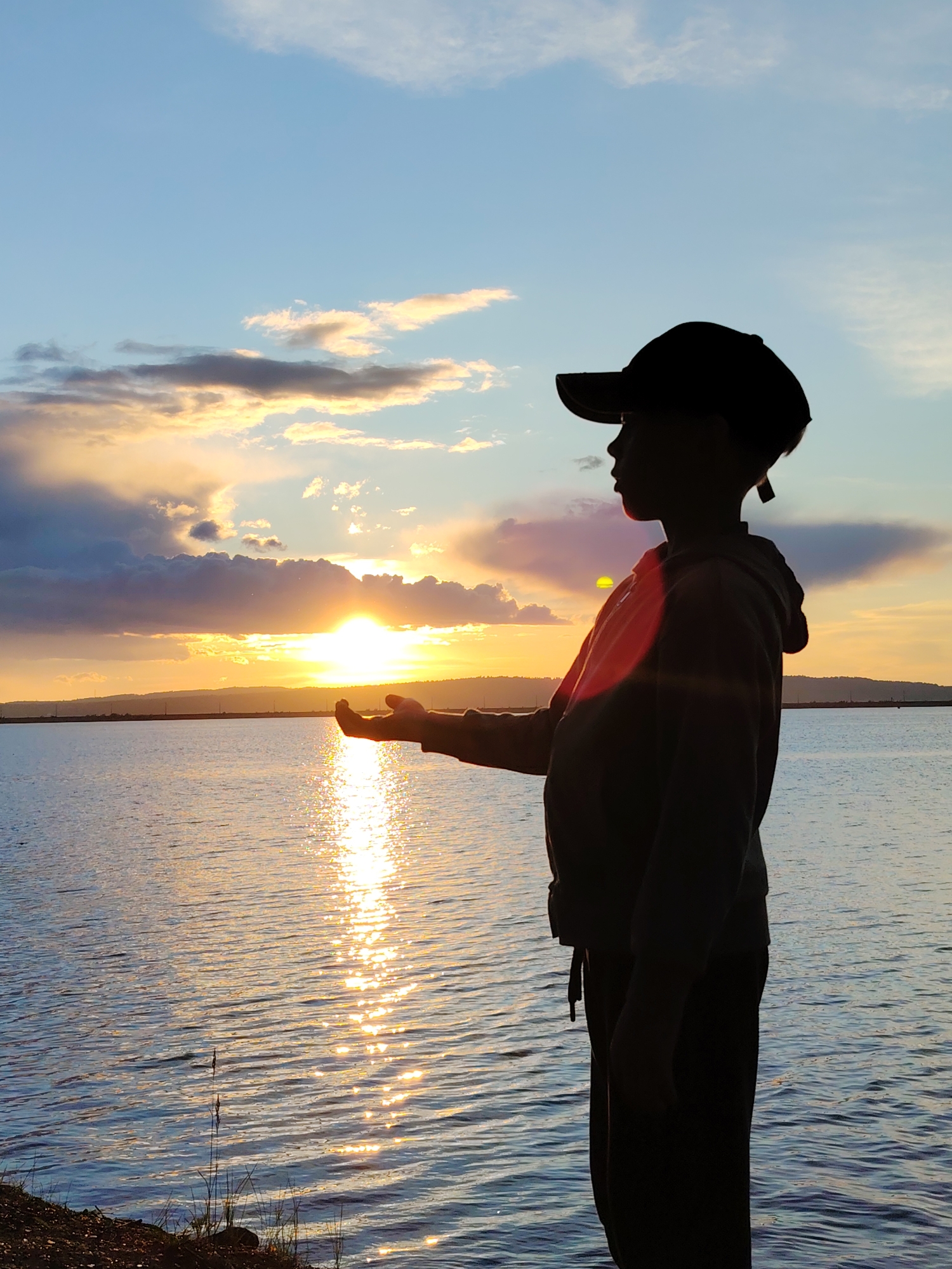 A young person, silhouetted against a sunset sky, stands near a body of water in the city of Chaikovsky, Russia. The child wears a cap and a hooded sweatshirt. The setting sun reflects off the water. The landscape in the background suggests a lake or reservoir with distant land.