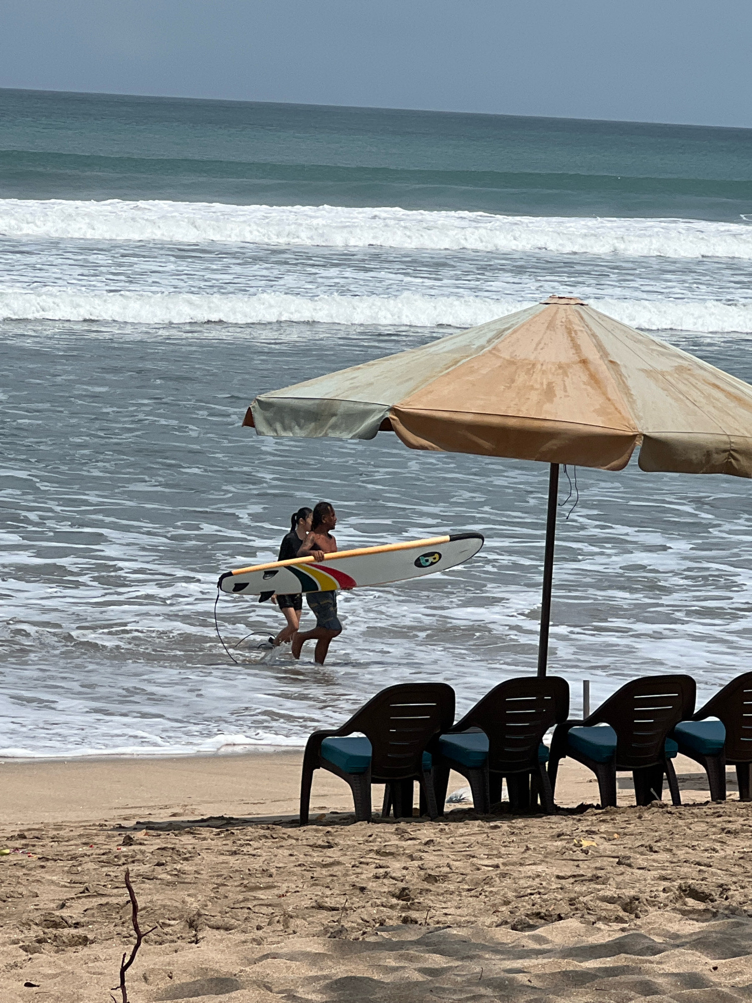 In Kuta, Indonesia, two individuals, a man and a woman, walk in the shallow ocean waves carrying a surfboard. The man, in patterned swim trunks, holds the surfboard horizontally with the woman, wearing a dark top and shorts, beside him. A large, neutral-colored beach umbrella stands on the sandy shore behind them, casting a shadow. In the foreground, several empty dark plastic beach chairs with teal cushions are arranged in a row. The ocean exhibits moderate waves with white foam, and the sky is overcast.