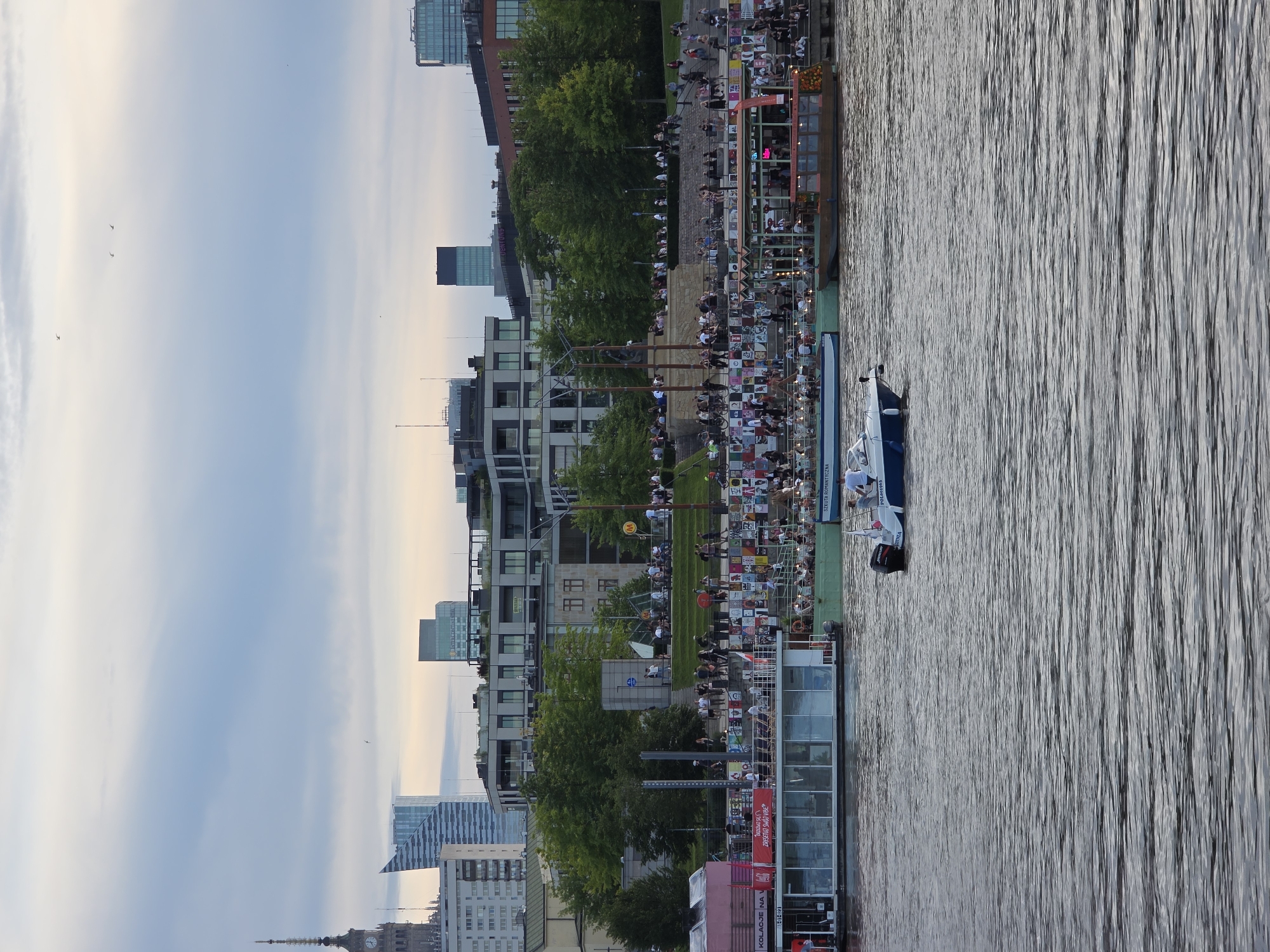 The image depicts the Vistula River waterfront in Warsaw, Poland, near the Copernicus Science Centre, under a cloudy sky. In the foreground, the water is textured with ripples. A white and blue motorboat, with two individuals visible, navigates the river. In the midground, a series of moored boats and platforms line the riverbank. One platform, a long, glass-enclosed structure, displays promotional materials and is filled with people. Adjacent to it is a larger, wooden boat with an open upper deck, also occupied by numerous individuals. The riverbank itself is tiered, with a wide promenade populated by a crowd of people, some seated on steps or benches, others standing and observing. Green trees and grassy areas are visible behind the promenade, interspersed with modern buildings and high-rise structures that characterize Warsaw's skyline. Notably, the Palace of Culture and Science's spire is visible in the distance, a landmark of the city. The overall scene suggests a lively public gathering and recreational activity along the river.