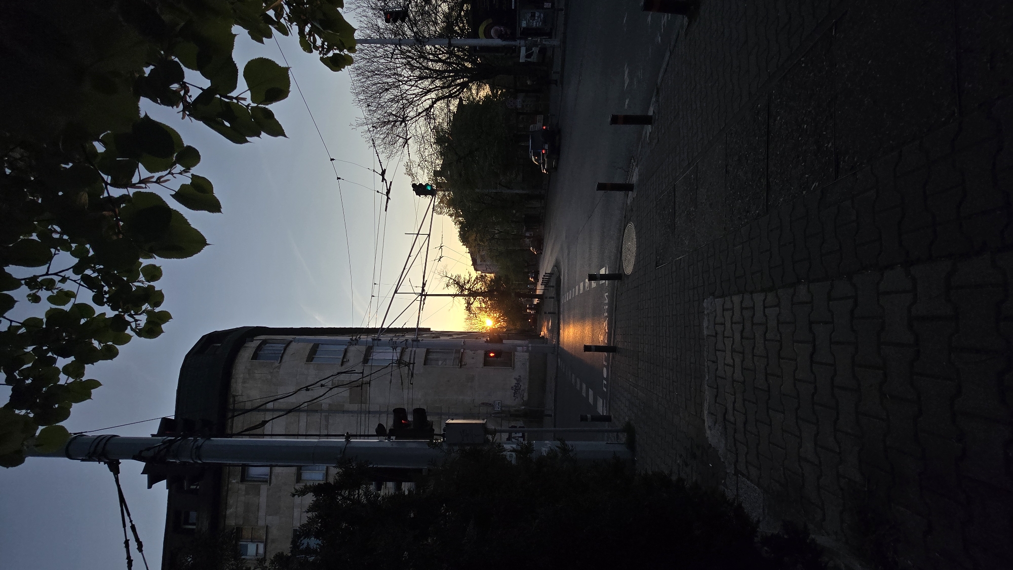 The image depicts an urban scene in Stara Zagora, Bulgaria, captured during sunset. The camera angle is low, looking upward towards the intersection of a street. A tall, light-colored building with a rounded corner occupies the left side of the frame. A utility pole is present in front of the building, with wires extending across the street.
Traffic lights are visible, both red and green, indicating their operational status. A car is present in the street, moving away from the viewer towards the sunset. The road is paved and reflects the orange light of the setting sun, creating a visual effect of illumination.
Sidewalks made of interlocking bricks are along the right side of the frame. Wooden bollards are spaced along the sidewalk edge. Trees and foliage frame the image on the left and top, some silhouetted against the bright sky. The sky is a gradient of blue to orange, indicative of the time of day.