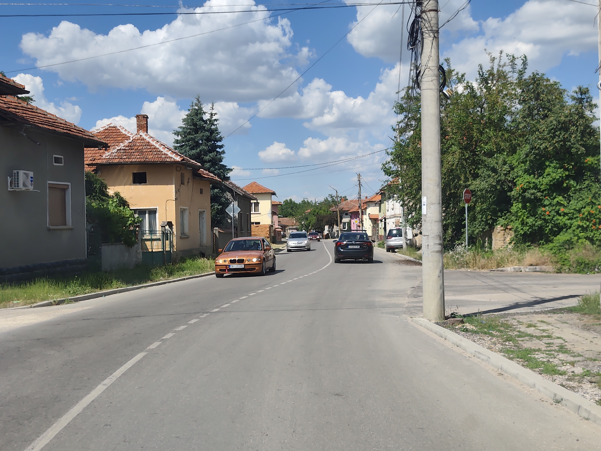 This image captures a sunny day scene on a street in Dermantsi, Bulgaria. The road curves to the right, lined with residential buildings on both sides. Houses with red-tiled roofs and beige or light-colored walls are visible. Some have air conditioning units mounted on their exterior. Lush green trees and grass flank the road, contributing to a peaceful, suburban atmosphere.
Several cars are on the road, with an orange sedan in the foreground on the left, a silver car further ahead, and a dark sedan and a silver hatchback in the distance. The activity appears to be typical daily traffic in a village or small town. The sky is bright blue with fluffy white clouds, suggesting pleasant weather, likely midday or early afternoon given the shadows cast. A prominent utility pole with numerous wires stands on the right side of the road, a common sight in many locales. A red stop sign is also visible on the right, indicating a potential intersection or hazard.
The overall impression is one of a quiet, lived-in community under a clear sky. There is no visible text in the image other than the license plate on the orange car which reads "CB 1193 BD".