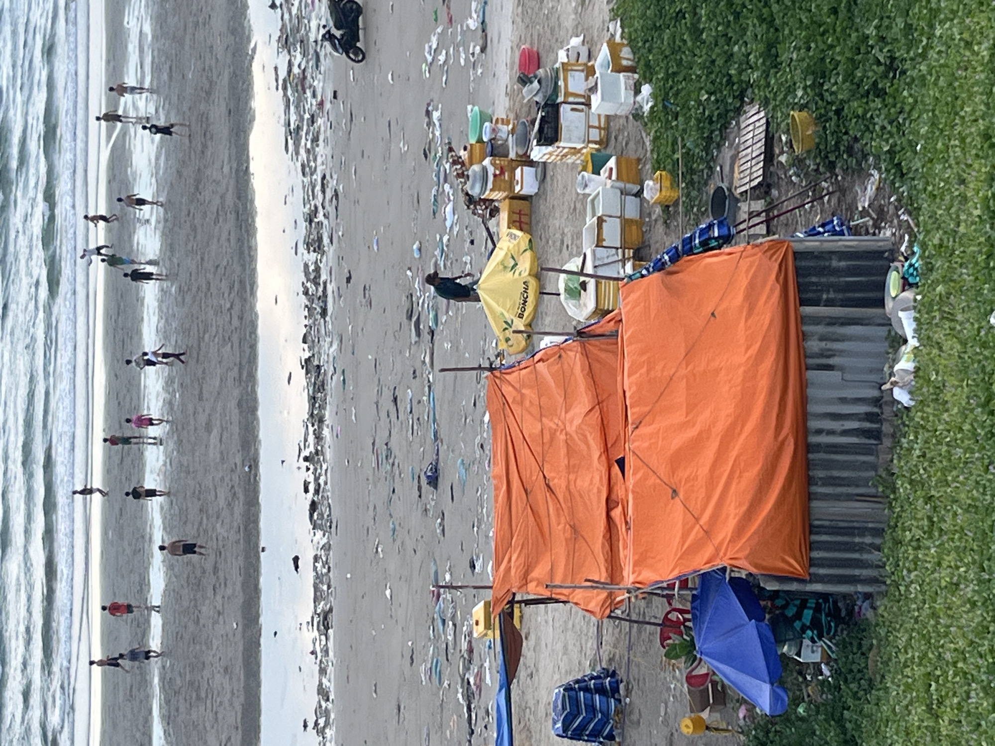 The image depicts a littered beach in Mũi Né, Vietnam. Numerous people are scattered across the sandy shore, some walking along the water's edge, others appearing to be playing or exercising. The ocean waves are visible on the left side of the frame. A significant amount of trash, including plastic bags and debris, is strewn across the beach, creating a stark contrast with the natural environment.
In the foreground, a makeshift stall or shelter is set up, covered by bright orange tarps. Various objects, possibly goods for sale, are stacked around the stall, including boxes, buckets, and containers. A yellow umbrella with visible text, "BONCHA," is also part of the stall's setup. A blue umbrella is positioned near the base of the stall. Behind the stall, a corrugated metal structure and lush green vegetation are visible, suggesting a more permanent presence or a boundary to the beach.
The overall impression is one of a busy beach environment marred by pollution. The presence of the stall indicates commercial activity, likely catering to beachgoers. The time of day appears to be daytime, with overcast or diffused lighting. The image captures a scene that highlights both human activity and the environmental challenges faced by popular tourist destinations.