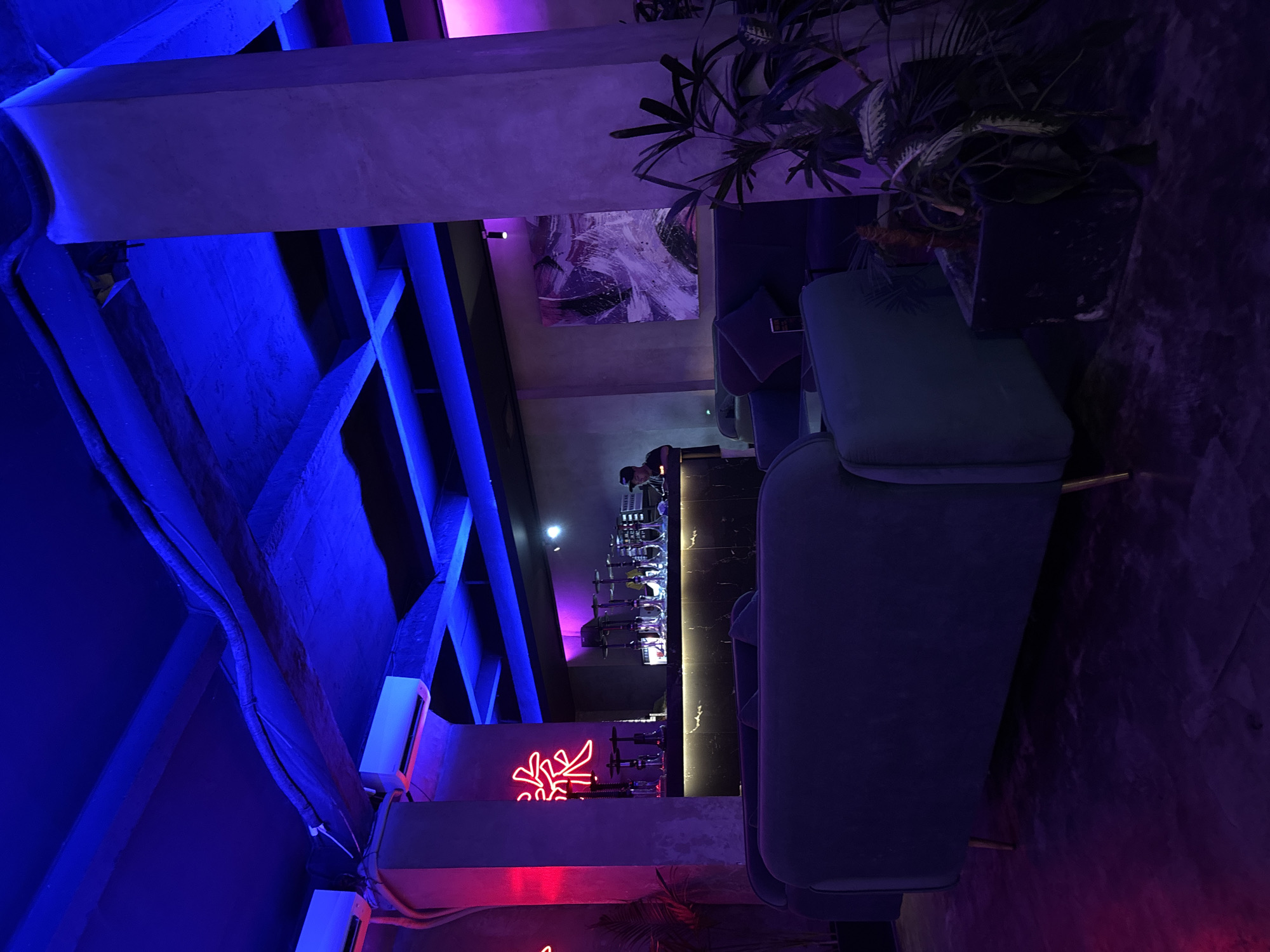 Captured inside what appears to be a bar or lounge in Kuta Selatan, Indonesia, the image presents a space bathed in blue and purple neon lighting. Concrete beams support the ceiling and a central bar is visible, partially obscured by modern furniture. A person is seen behind the bar, presumably a bartender. Neon signs decorate the walls, while potted plants add a touch of nature to the scene. The image is taken from an unusual angle.
