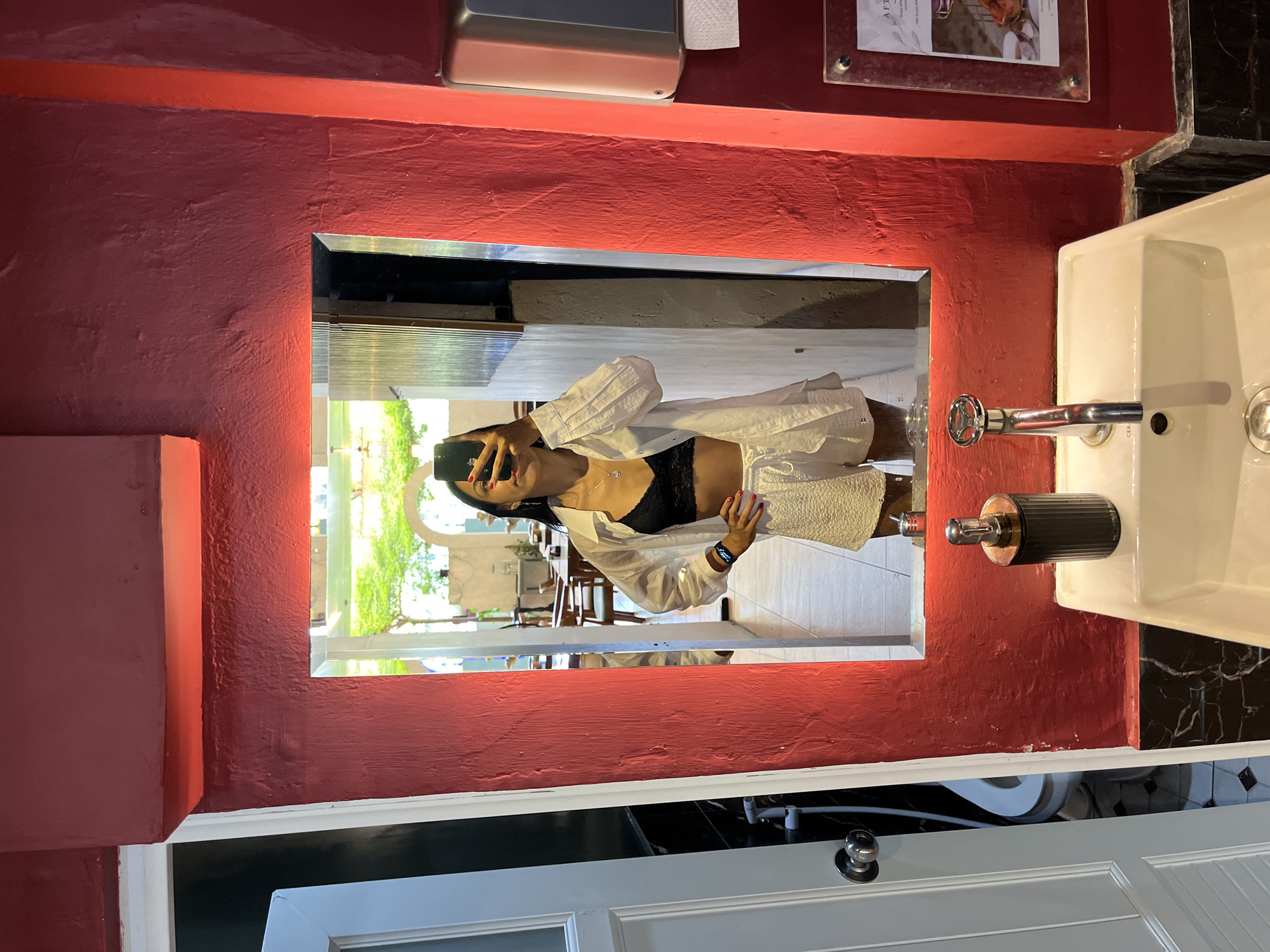 A woman stands in a bathroom in Kuta Selatan, Indonesia, taking a mirror selfie with a smartphone. The mirror is framed by a thick, textured red painted wall. The bathroom features a white sink with silver fixtures, a black soap dispenser, and a partially visible white door with a silver knob. The reflection in the mirror reveals an outdoor dining area with tables and chairs, greenery, and natural light. The woman is wearing a black lace top and a white button-down shirt. A framed sign is above the mirror. The wall adjacent to the sink has black marble elements.
