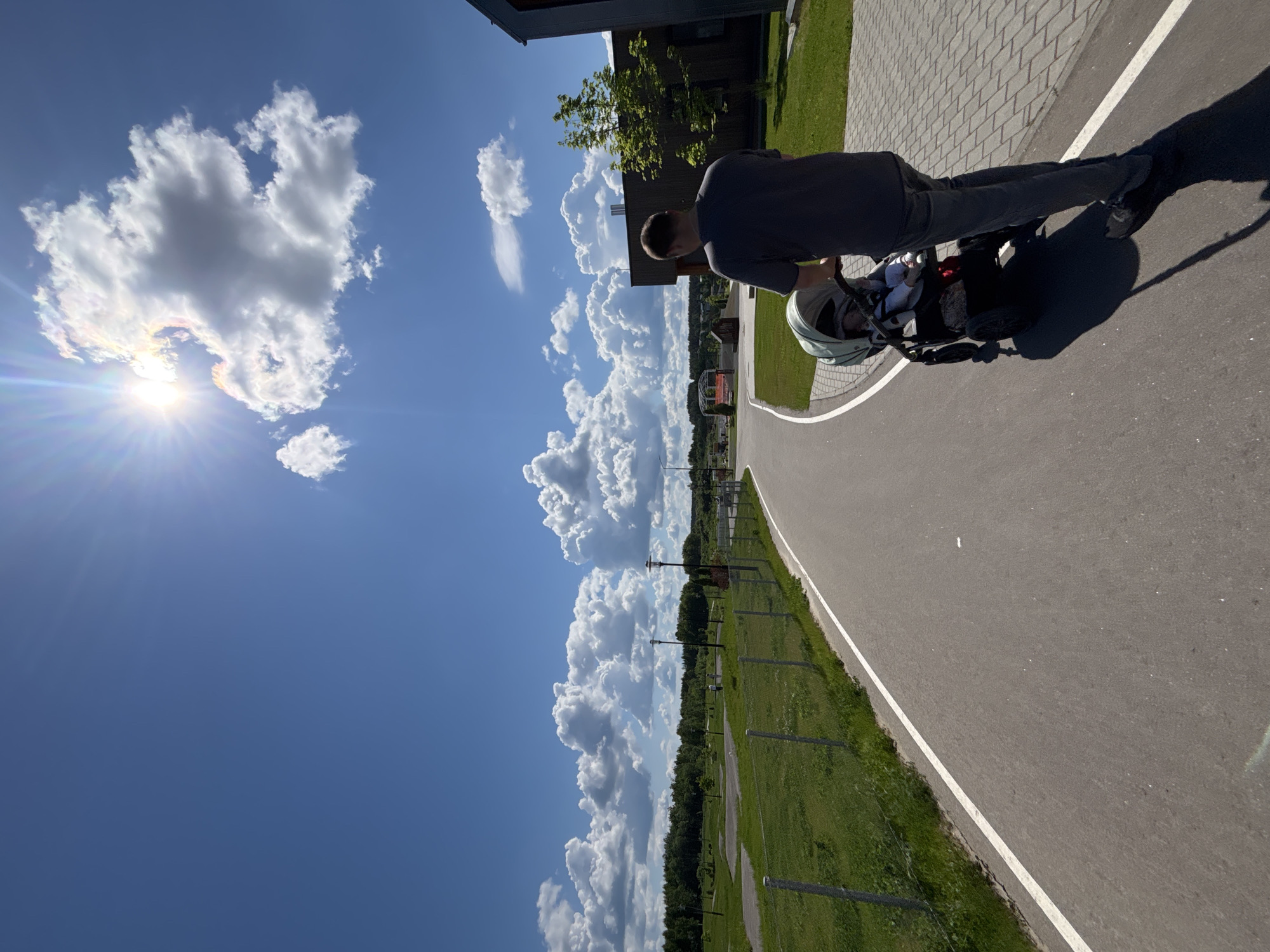 A man in a gray shirt and dark pants stands on a gray asphalt path in Varaksino, Russia. He is pushing a light gray baby stroller. The path has a white line along one side, bordering a green grassy area. In the distance, a line of trees and possibly power lines is visible against a backdrop of fluffy white clouds in a blue sky. The sun is shining brightly, creating a lens flare effect in the top left corner of the image. The shadow of the man and stroller is cast on the path. A building is partially visible in the top right corner, with green grass surrounding it.