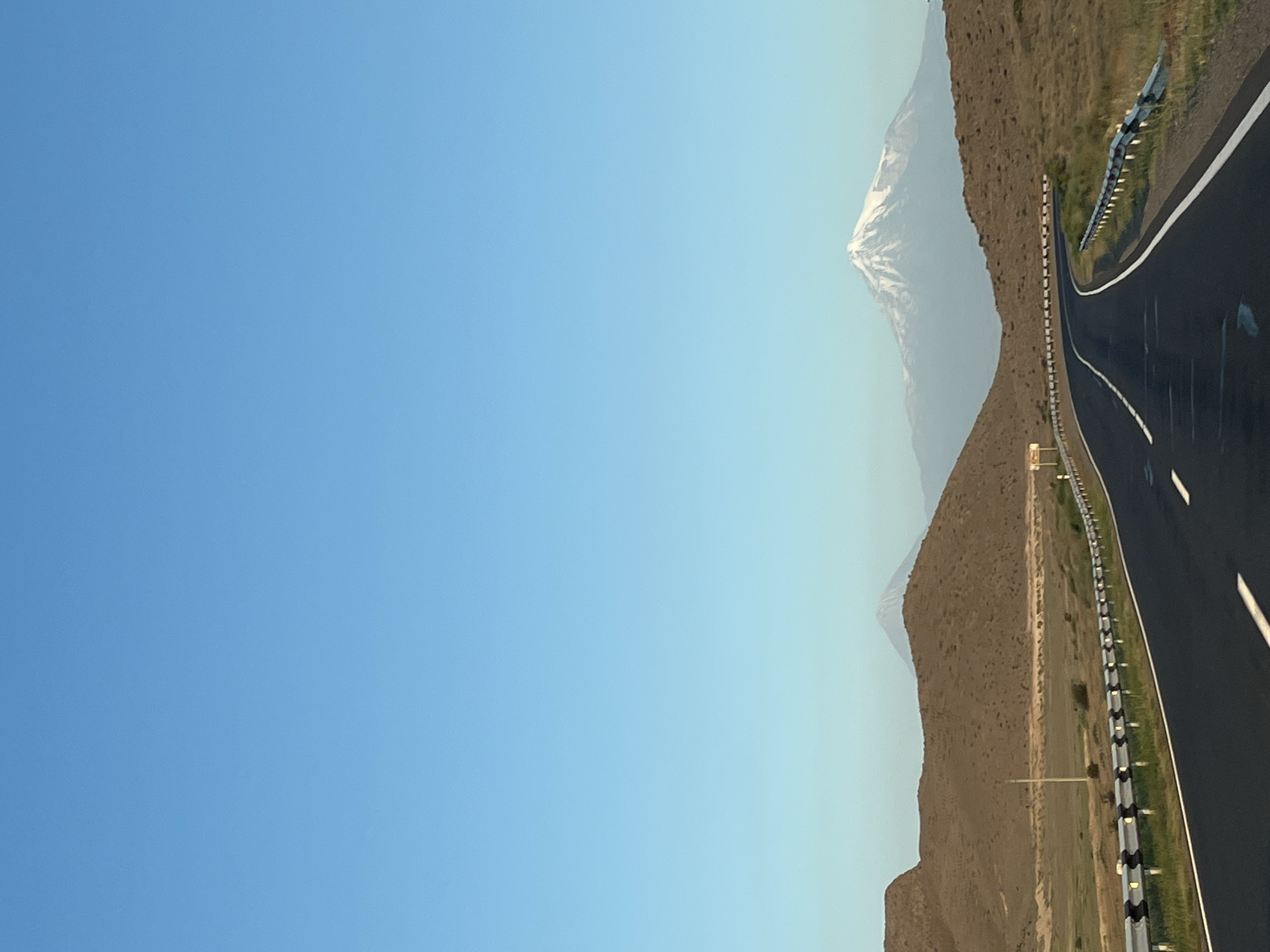 The image shows a highway curving through a barren, hilly landscape with mountains in the distance. The sky is clear and blue. In the foreground, a black asphalt road is bordered by a guardrail. The hills are covered in dry grass and scrub. In the background, two mountains are visible, one of which is snow-capped. Based on the visual cues, this scene is likely Mount Aragats, a prominent mountain in Armenia, and the highway is likely a road leading to or from it. Given Zangakatun's proximity to Mount Aragats, this scene could be located in the vicinity of the city. There are no people or animals visible in the image.