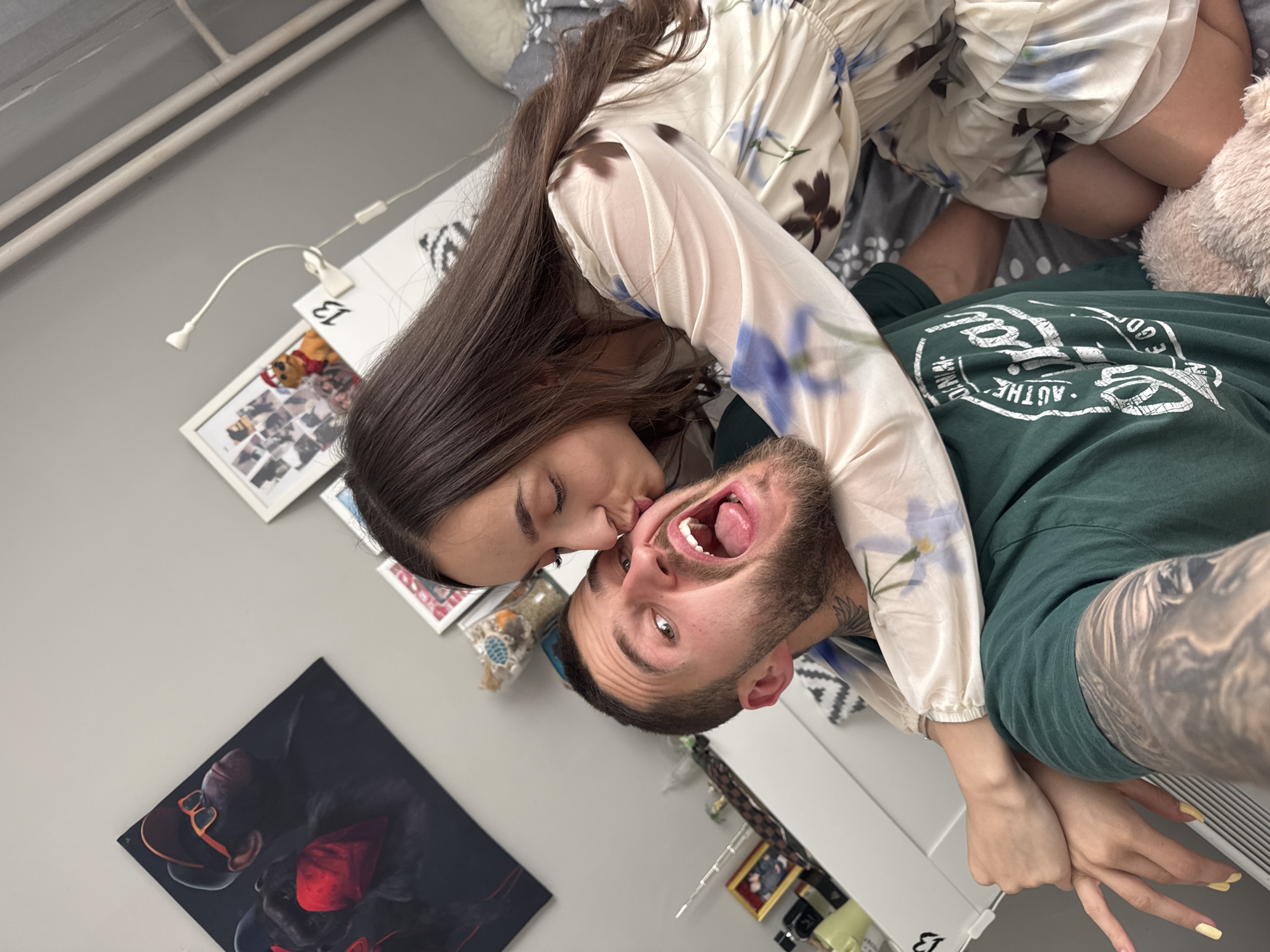 A couple lies on a bed in an interior setting. The woman, with long brown hair and wearing a floral-patterned top, kisses the cheek of the man, who is wearing a green t-shirt and making a silly face. The room has white furniture and decorations, including framed pictures on the wall. The location appears to be indoors, potentially a bedroom or living space. I cannot place this specific location in the city of Sofia, Bulgaria.