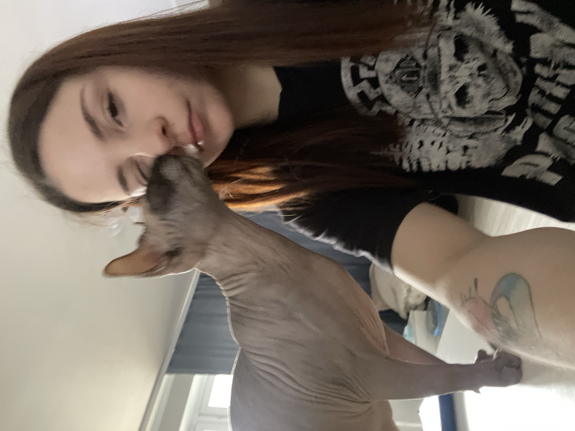 A woman is pictured in a selfie, tilted to the left. She has long brown hair and is wearing a black t-shirt with a skull design. A grey hairless cat is positioned close to her face, near her nose. The woman has a tattoo on her arm. The background shows a window and part of a room with light-colored walls. The location appears to be indoors.