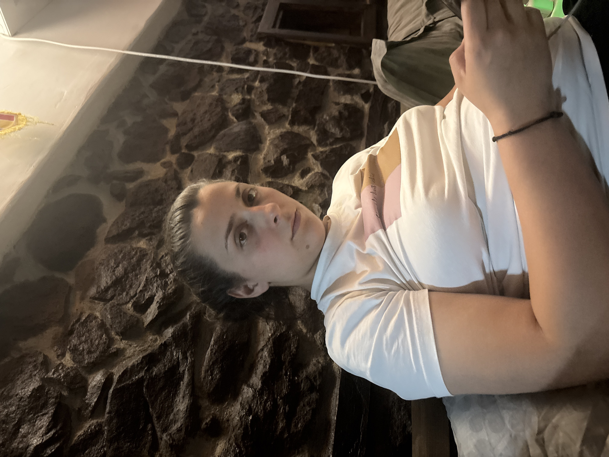 A woman is centrally featured in an indoor setting, positioned on the right side of the frame and captured from the chest up. She has dark hair pulled back and looks directly at the viewer with a calm, neutral expression; a small mole is visible on her left cheek. She wears a white, short-sleeved t-shirt, beneath which a pink and light brown garment with faint, light-colored text is visible on her right shoulder area, appearing to read "first rule." A thin, dark bracelet is on her right wrist. Her left arm is bent with her hand near her chest, while her right arm rests on a light-colored, patterned surface, likely upholstery.
The background is dominated by a dark, rustic stone wall, composed of irregularly shaped dark grey and brown stones with lighter mortar lines. In the upper left, a white ledge or shelf holds a small, yellow and red decorative item. A thin white cable runs horizontally across the upper wall. To the upper right, a dark, rectangular opening, possibly a window or niche, and some draped olive green or dark fabric are visible.
The scene depicts the woman posing for a photograph. The lighting is dim and diffused, suggesting an indoor evening environment. The rustic stone wall and potential traditional decor hint at a characteristic interior in Belopoptsi, Bulgaria.