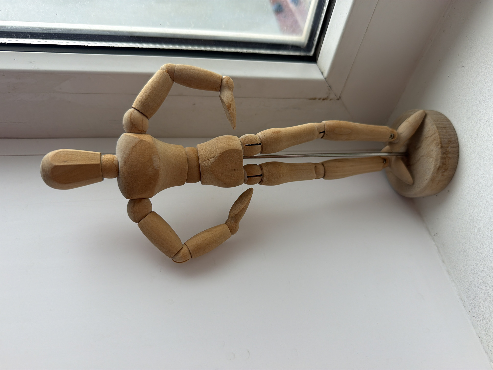 A wooden articulated artist's mannequin stands on a round wooden base on a white windowsill. The mannequin is positioned facing forward, with its arms bent and hands resting on its hips. It is made of light-colored wood and has visible joints. Behind the mannequin is a white wall. To the right of the mannequin is a window with a dark frame. The window looks out onto an indistinct outdoor scene. There are no people present in the image. This scene is not specifically identifiable as being in Yuzhno-Sakhalinsk, Russia, or any other specific city.
