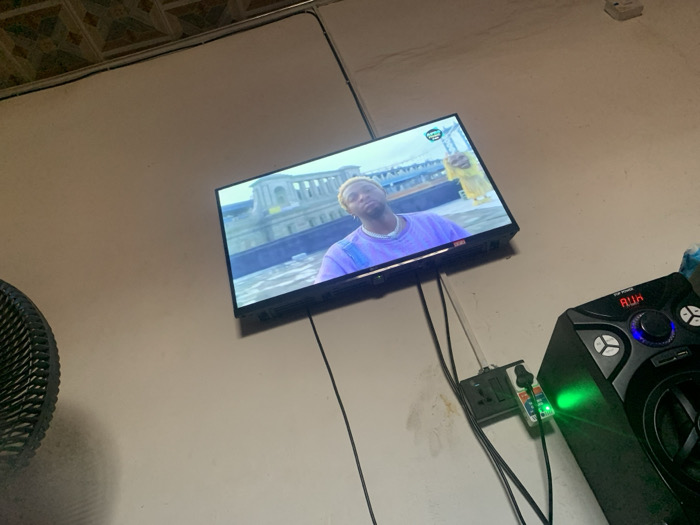 A television is mounted on a white wall displaying a man wearing a purple shirt and a silver chain, holding a yellow bottle. Below the TV, electrical cords run to a power strip. A black speaker with multiple buttons and a green light is positioned to the right of the power strip. The room has a visible ceiling with a patterned tile design and an electric fan is partially visible on the left side.