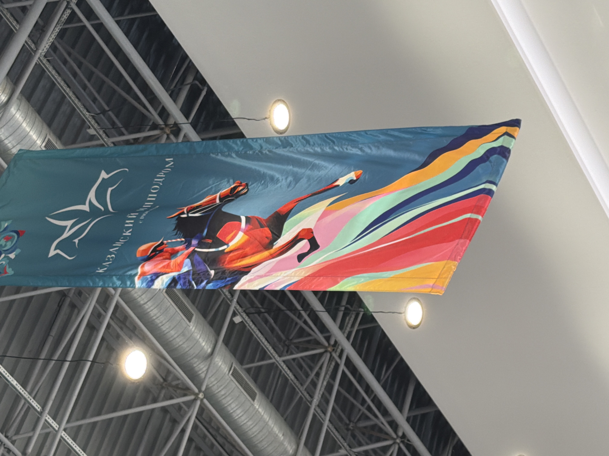 This media file displays a banner with a vibrant graphic of a horse in motion, depicted with a stylized, abstract design. The horse is shown with its legs extended, suggesting speed and power, and is rendered in bold colors such as red, orange, yellow, blue, and green, forming a dynamic, wave-like pattern that trails behind it. The banner is predominantly teal on the left side, featuring a white silhouette of a bird, possibly a falcon or eagle, with its wings spread.
Text is visible on the teal portion of the banner in white Cyrillic letters. The visible text appears to be "КАЗАХСКИЙ ИПОДРОМ" which translates to "Kazakhstan Hippodrome" or "Kazakh Hippodrome". Below this, there's smaller text that is partially obscured.
The banner is suspended indoors, with a complex industrial ceiling structure visible above it, composed of metal beams and pipes, likely in a large hall or stadium. Circular ceiling lights are also visible, contributing to the overall illumination of the scene. The lighting suggests an indoor event or exhibition space. The image is taken from a low angle, looking upwards at the banner and ceiling. There are no people clearly visible in the frame, and the focus is on the banner and its artistic design. The overall impression is one of dynamism and perhaps a celebration of equestrian sports or a related event.