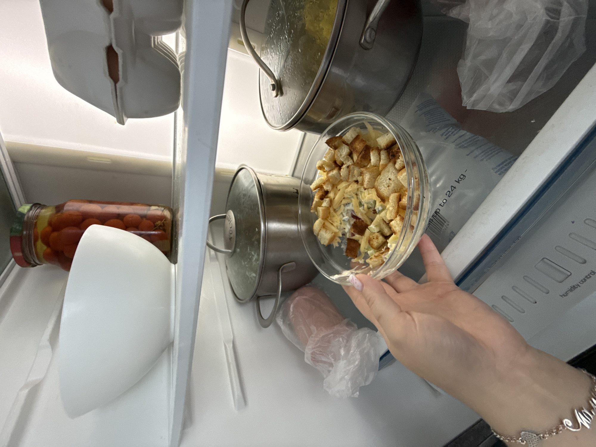 A person's left hand, adorned with a bracelet and nail polish, holds a clear glass bowl filled with croutons and shredded cheese. The bowl is positioned in the foreground, on a shelf inside a refrigerator. Behind the bowl, on the same shelf, are two stainless steel pots with lids. To the left of the pots, a pink item is wrapped in clear plastic. On the shelf above, a large white bowl is visible, and to its right, a glass jar filled with red cherry tomatoes. Further back, an egg carton containing brown eggs is present. The refrigerator interior is white. The location is the city of Stary Oskol, Russia.