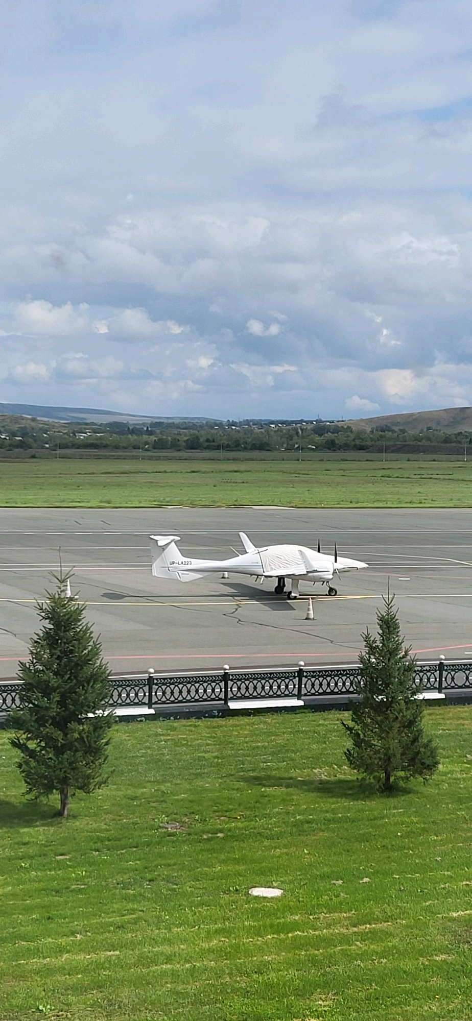 The image shows a twin-engine airplane with the registration UP-LA223 parked on an asphalt runway at the Oskemen Airport in Oskemen, Kazakhstan. The airplane is white and has a tarp covering its cockpit and front section. Two conical orange markers are placed near the airplane. In the foreground, there are two evergreen trees and a patch of green grass. Behind the runway, there is a decorative fence and a vast expanse of green fields stretching towards rolling hills covered in trees and some buildings, indicating a rural or semi-rural setting on the outskirts of the city. The sky is overcast with scattered clouds.