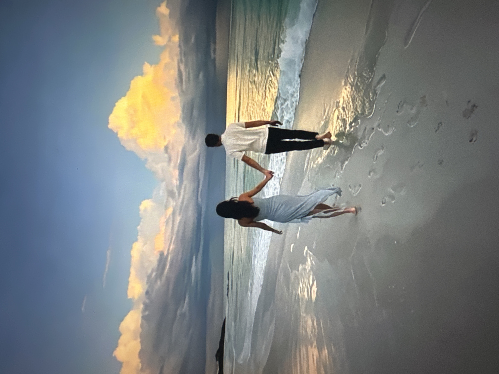 A couple walks hand-in-hand on a beach at sunset. The woman, in a light blue dress, walks in front of the man, who wears a white polo shirt and dark pants. Their footprints are visible in the wet sand. The ocean is to their right, with waves gently rolling onto the shore. The sky is a dramatic mix of blue, gray, and vibrant orange clouds, illuminated by the setting sun. The location is described as Unknown, Unknown.