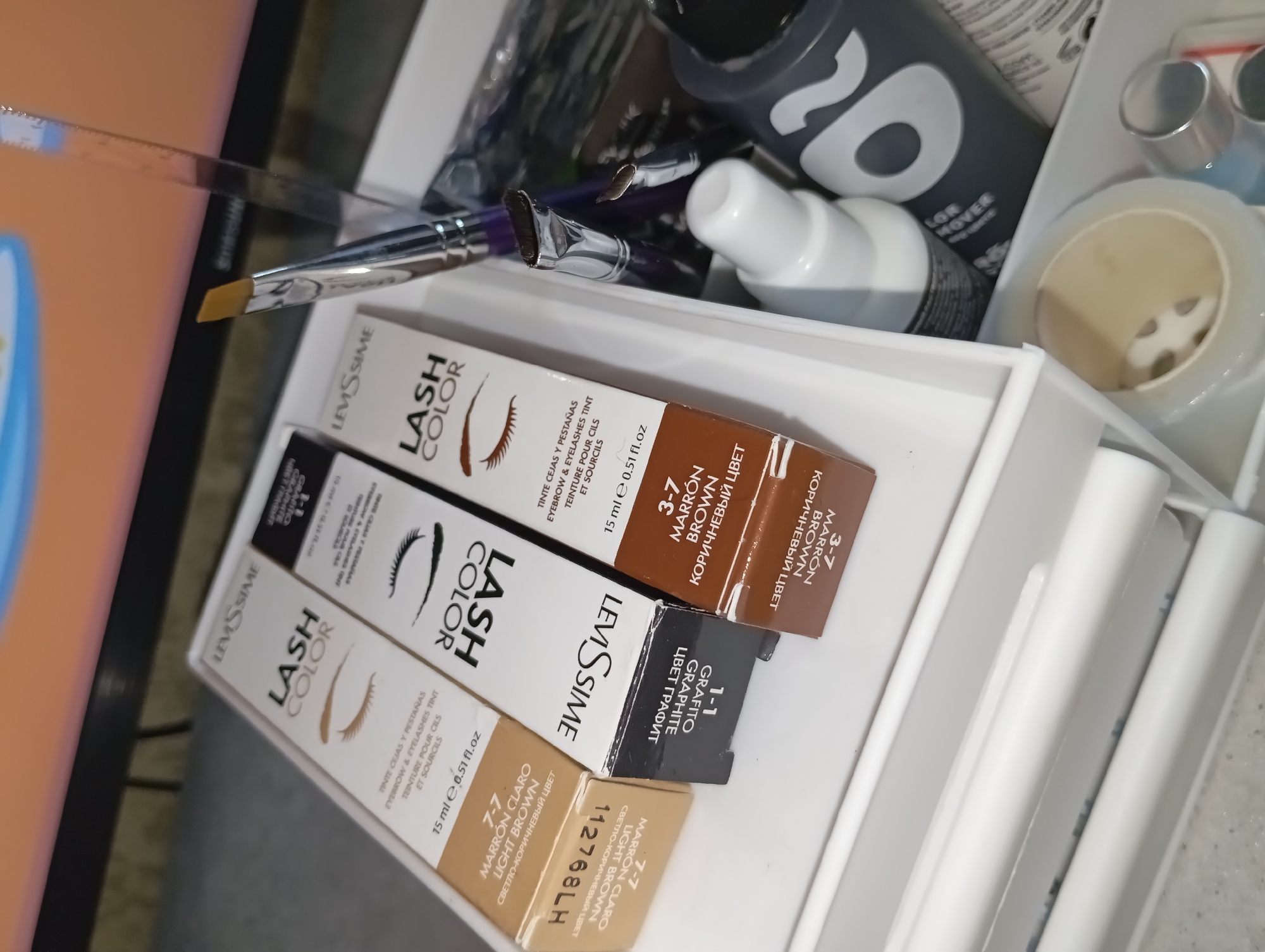 A still-life composition features a white storage drawer filled with beauty and cosmetic supplies, potentially located in Yurgamysh, Russia. Dominating the scene are three LevisSime Lash Color boxes in shades of Light Brown (7-7), Graphite (1-1), and Brown (3-7). Adjacent to the Lash Color boxes is a makeup brush with a silver handle and tan bristles, lying across other cosmetic brushes. Several bottles are also present in the drawer, including a white squeeze bottle and a gray bottle with a large "20" on the front. A roll of clear tape and other cosmetic items fill the remaining space.