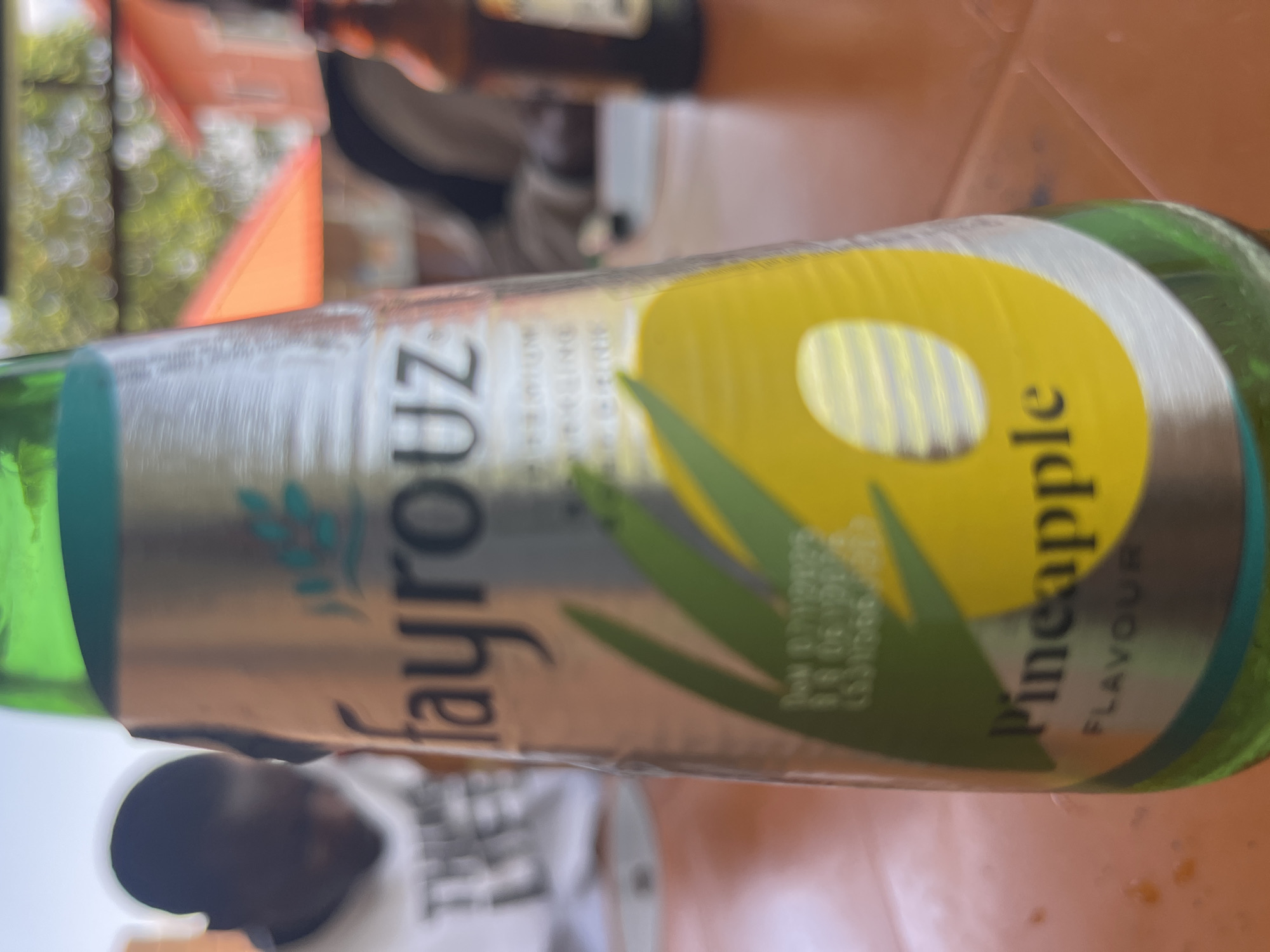 The image shows a close-up of a Fayrouz Pineapple flavour soda bottle on an orange table, with an out-of-focus background. In the background, a person in a white t-shirt is visible on the left, and another person is seated on the right, with a brown glass bottle next to them. Buildings and trees are also discernible in the distant background. The overall setting suggests an outdoor dining or relaxation area. The specific location in Jos, Nigeria is not evident from the image alone.