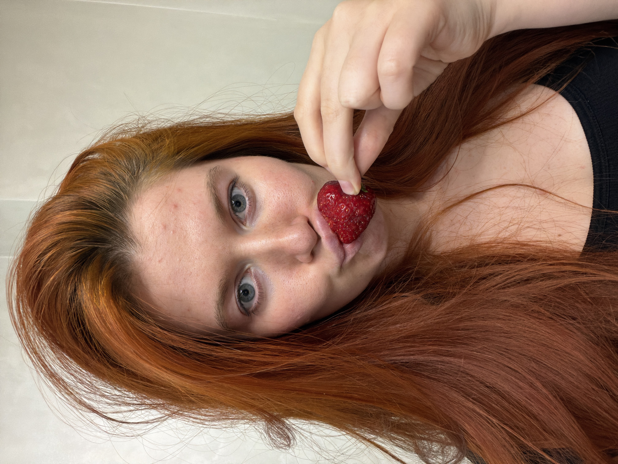 A woman with long, red hair and blue eyes is holding a strawberry to her lips, with her mouth puckered. She is wearing a black top. The background is a plain, light-colored wall. The scene appears to be an indoor setting, possibly a bathroom or bedroom, given the plain wall and lighting. There is no discernible indication of the location being in Krasnodar, Russia.