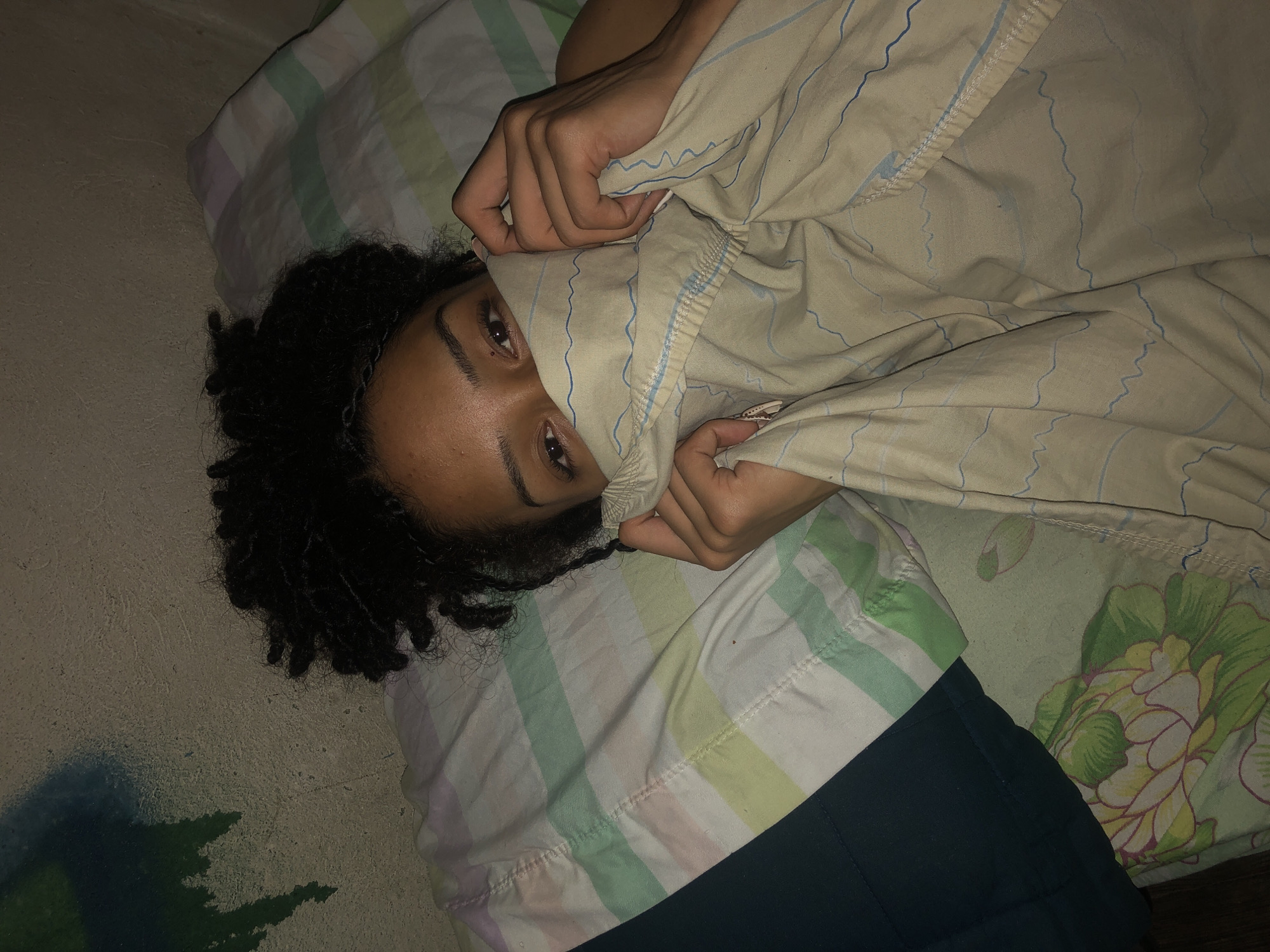 A person with dark curly hair is lying on a bed. Their eyes are visible above a tan-colored sheet with blue wavy lines that they are holding up to their face with both hands. The bed has striped and floral patterned bedding. The room appears dimly lit. The setting lacks distinctive Argentinian details. No evidence indicates the location is Buenos Aires.