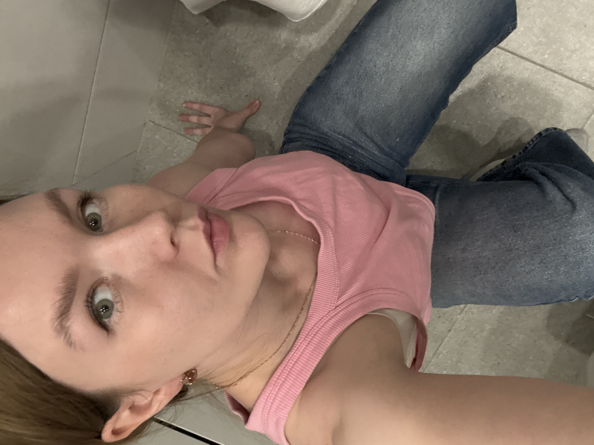 A young woman is sitting on the floor indoors. She has light skin, light brown hair, and green eyes. She is wearing a pink tank top and blue jeans. Her left hand is resting on the floor next to her. The floor appears to be tiled. There are no discernible elements in the image that would suggest a specific location in the city of Izhevsk, Russia, or any other specific city. The environment appears to be a domestic or public indoor space.