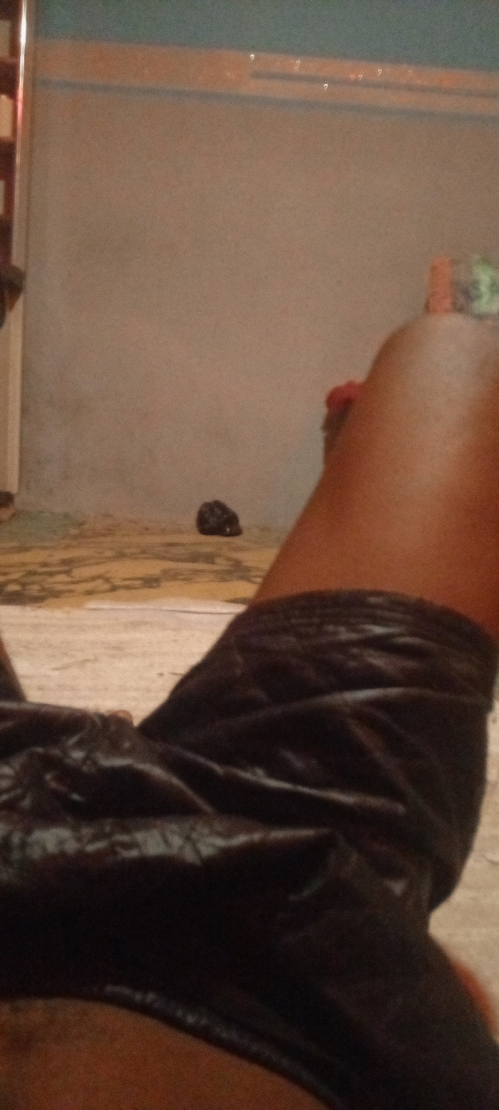 The image presents an indoor scene from a first-person perspective, looking down at a person's legs while they are resting or reclining. The subject's bare legs, which have a dark brown skin tone, are visible, extending into a pair of dark, possibly black, shorts. The shorts appear to be made of a soft, slightly textured or velvety material.
In the background, a light-colored floor with abstract, darker patterns is visible, suggesting either patterned tiles or a rug. Further back, a plain, light grey or beige wall rises, topped by a darker horizontal trim line. Above this trim, the upper section of the wall appears in a muted blue-green hue. Several small, faint, and indistinct reflective marks, which in close-up resemble symbols or inverted question marks, are sparsely visible on this upper wall area. On the floor, closer to the main wall, a small, dark, unidentifiable object is present.
The lighting in the scene is dim and warm-toned, suggesting low natural light or artificial illumination, possibly during the evening or night. The overall impression is one of casual relaxation. The location context is Ago-Abo, Nigeria.