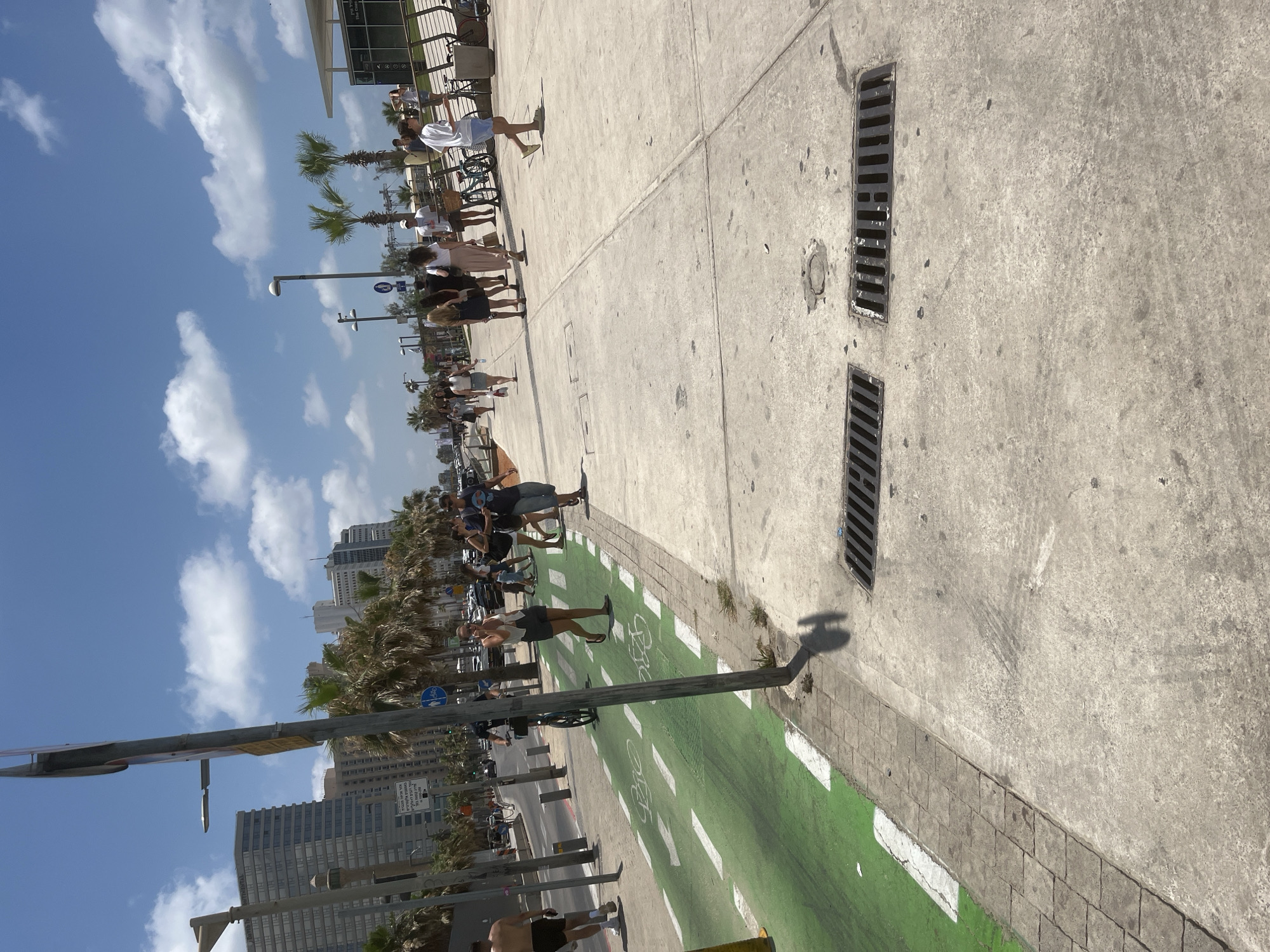 The image shows a sunny day with scattered clouds at a beach promenade in Tel Aviv, Israel. In the foreground, a concrete sidewalk with two grates runs alongside a bright green bicycle lane marked with white bicycle symbols. People are walking on the sidewalk. In the background, there is a street with cars and palm trees. Tall buildings line the street, suggesting an urban environment. The overall scene depicts a lively public space designed for recreation and urban living.