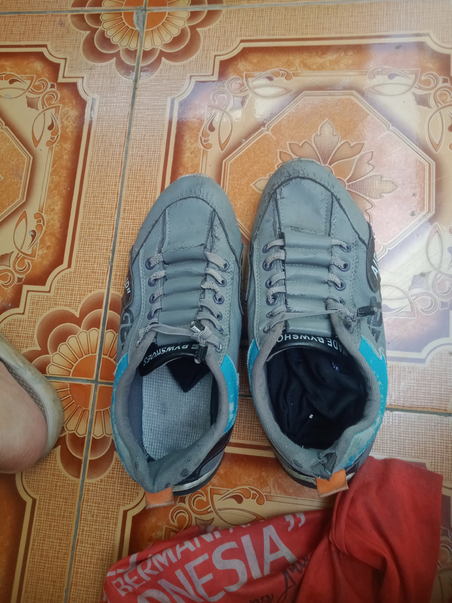 Two gray athletic shoes are placed side-by-side on a patterned tiled floor. The shoes are laced with gray laces and have blue accents around the collar. One shoe is slightly closer to the camera and its sole is visible. To the left of the shoes, a portion of a light-colored shoe is visible. To the lower right, a red garment with white lettering, possibly a t-shirt, is partially covering the floor. The tiled floor has an orange and brown geometric pattern with floral motifs. The image appears to be taken indoors, in a residential setting. The location is described as the city of Sumedang, Indonesia.