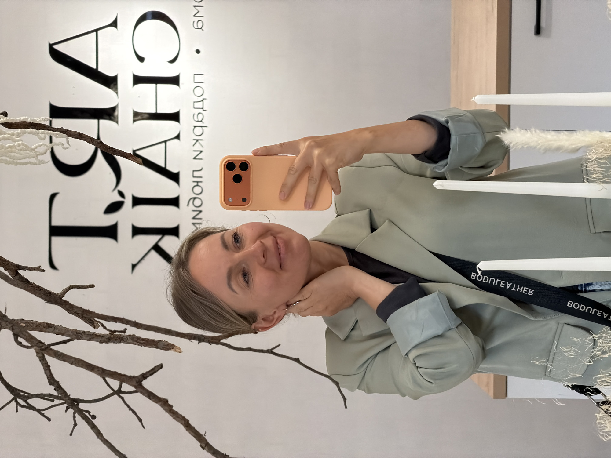The image, rotated 90 degrees clockwise, is a mirror selfie taken by a woman in Chaikovsky, Russia. She is smiling and holding a peach/orange-cased smartphone to capture her reflection. The woman has light brown hair and is wearing a light sage-green blazer over a darker top, with the blazer's sleeves cuffed.
The background is a white wall with prominent, dark Cyrillic text. Partially obscured by the woman and decorative elements, the text on the wall reads "АЙЯ ЧАЙ • ПОДАРКИ МОР". This translates to "AYYA TEA • SEA/MANY GIFTS," suggesting the location is likely a tea or gift shop. Natural, bare branches with white fluffy accents serve as decor in the foreground and midground. To the right, several white taper candles and more white feathery decorative pieces are visible.
A black strap, likely from a bag or garment, crosses the woman's torso, displaying mirrored white text that appears as a brand name. The overall setting is indoors, featuring a clean and minimalist aesthetic with natural elements. The woman's expression is happy and content.