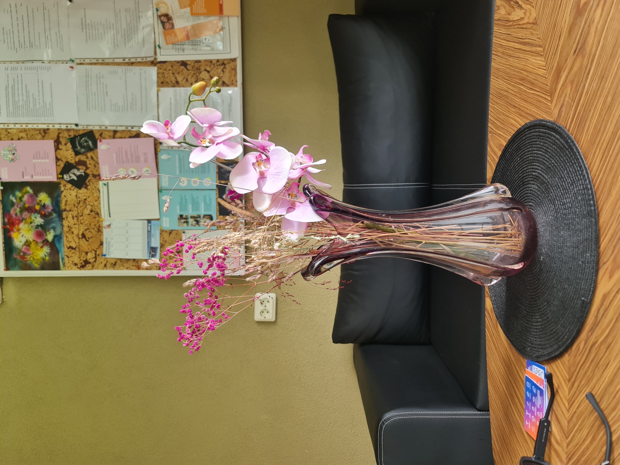 In a room with a textured olive-green wall, a black leather sofa is visible. On a wooden table in front of the sofa, a purple glass vase holds a bouquet of pink orchids and dried flowers. To the left of the vase, a black pair of glasses lies on the table next to a stack of three colorful cards. Behind the sofa, a bulletin board displays various papers, including a colorful floral picture at the top. A white electrical outlet with two sockets is mounted on the wall to the left of the bulletin board. The scene appears to be in a domestic or office setting in Molėtai, Lithuania.