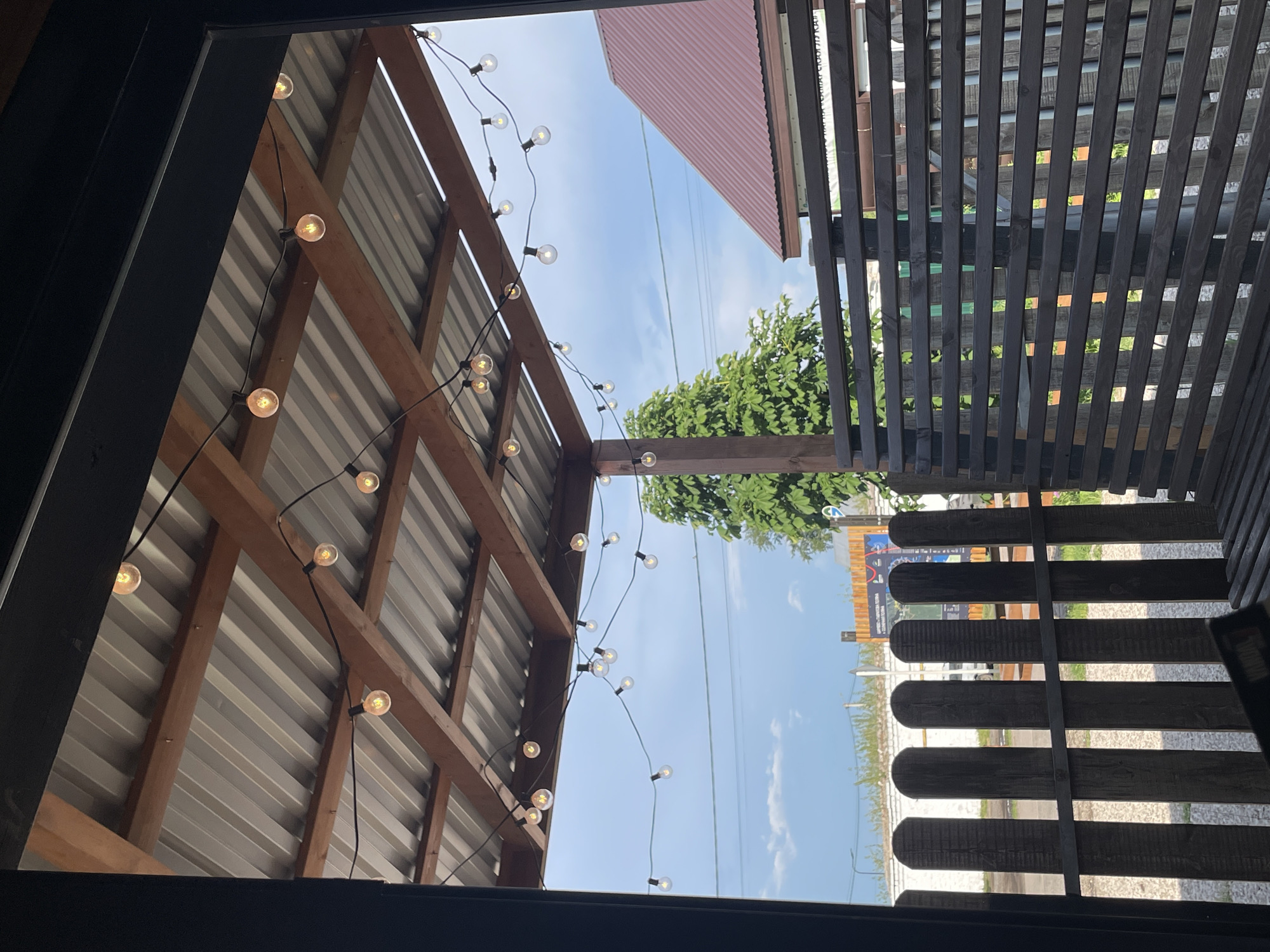 The image shows an outdoor area with a covered patio. The patio has a corrugated metal roof supported by wooden beams. A string of incandescent bulbs is strung across the underside of the roof. Through the opening of the patio, a wooden fence with vertical planks is visible, followed by a darker horizontal slatted fence. Beyond the fences, a large green tree stands to the left, and a building with a reddish-brown, corrugated roof is seen to the right. The sky is cloudy. In the background, there appears to be some kind of informational signage or display. The scene is consistent with an outdoor seating area at a restaurant or cafe in Shiryaevo, Russia, likely during daylight hours. There are no people or animals visible in the image.