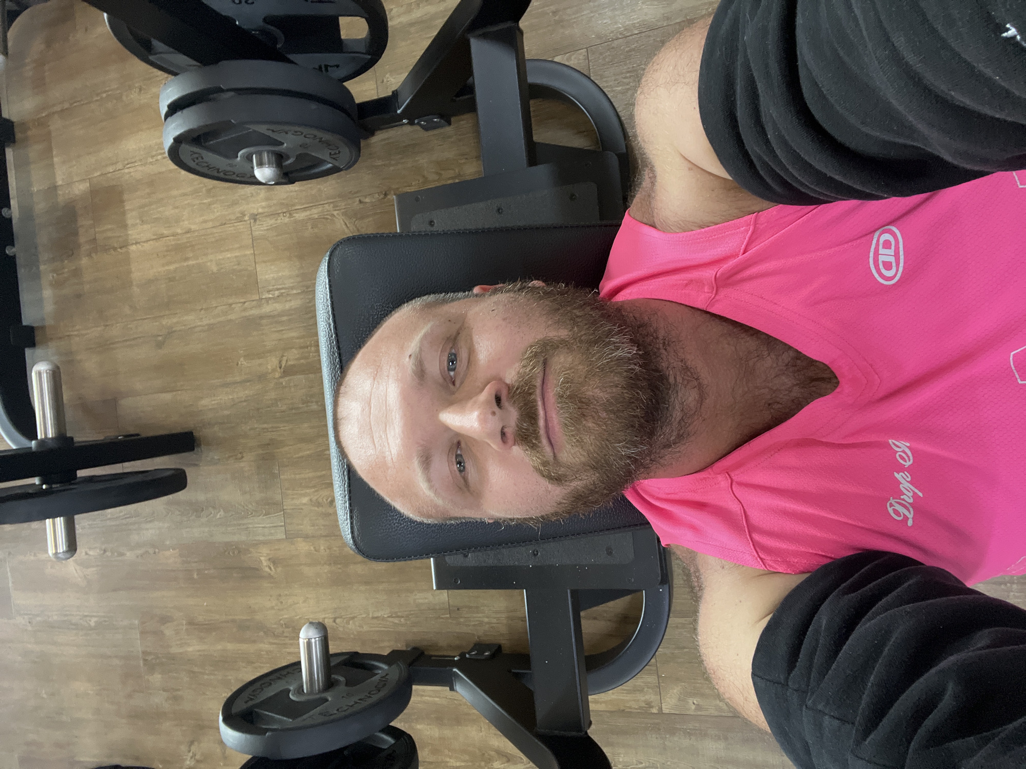 The image depicts a man with a beard lying on a weight machine in a gym. He is wearing a bright pink tank top and black arm sleeves. The gym has wooden flooring and weight plates are visible on the machine. The scene is shot from a high-angle perspective, looking down at the man. The image does not contain enough information to determine the specific location in Chaikovsky, Russia.