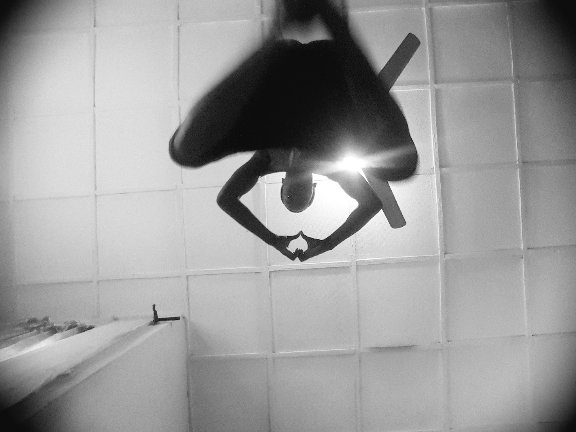 A black and white, low-angle, wide shot shows a person silhouetted against a bright light source, seemingly suspended from a ceiling that appears to be tiled or paneled. The person is in a contorted pose, with their body angled and limbs extended. One arm reaches up and to the left, while the other is bent at the elbow and extended downwards. Their legs are also positioned in a dynamic, acrobatic manner. The light source is positioned above and behind the person's head, creating a lens flare and obscuring some details of their face. The ceiling's grid pattern is visible throughout the image. The overall impression is one of athleticism and fluidity, captured in a moment of suspended motion. The specific location within the city of Unknown, Unknown cannot be determined from the image.