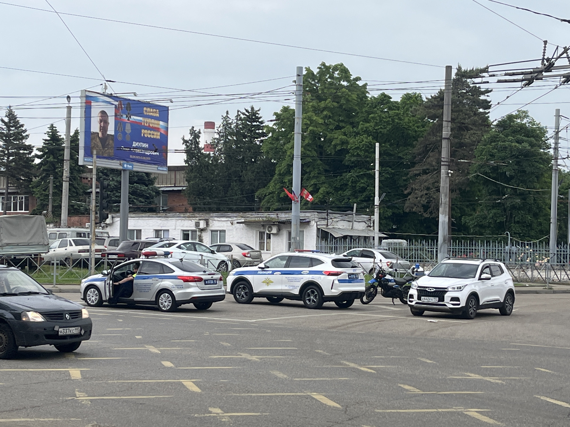 The image shows a street scene in Krasnodar, Russia. Three police vehicles are parked on the road. A black car is driving in the left of the frame, license plate "H337KC193". There's a motorcycle on the ground next to a white car, license plate "X442PX193". The license plate "M67283" is visible on the silver police vehicle with its door open. A billboard shows a portrait of someone labeled "ДЮЛИН" and text "СЛАВА ГЕРОЯМ РОССИИ." Green trees are in the background. Also in the background is a white building. A pole holds two red flags. Overhead wires crisscross the sky.