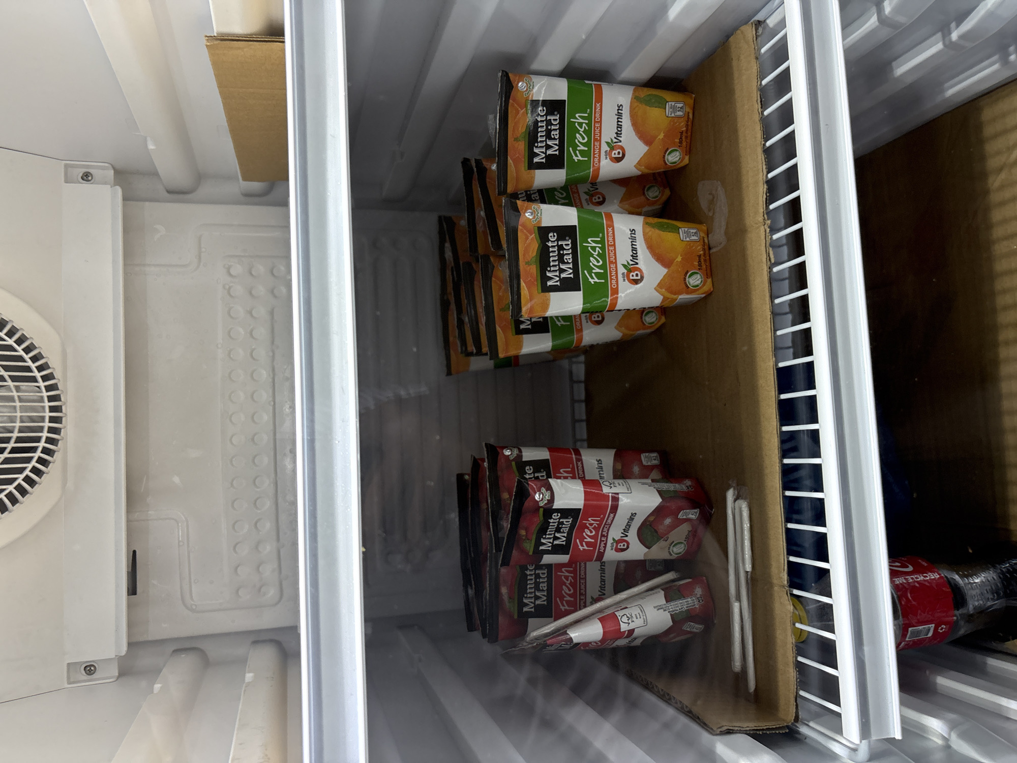 The image shows the inside of a refrigerator. On the top shelf, there is a cardboard box and a white plastic structure with a fan grille. The middle shelf contains several cartons of Minute Maid "Fresh" juice drinks, with two types visible: apple juice drink and orange juice drink. The apple juice cartons are on the left side, and the orange juice cartons are on the right. Several straws are placed on top of the cardboard base in front of the juice cartons. On the bottom shelf, a red plastic bottle, possibly a Coca-Cola product, is visible. The refrigerator appears to be in a domestic or commercial setting.