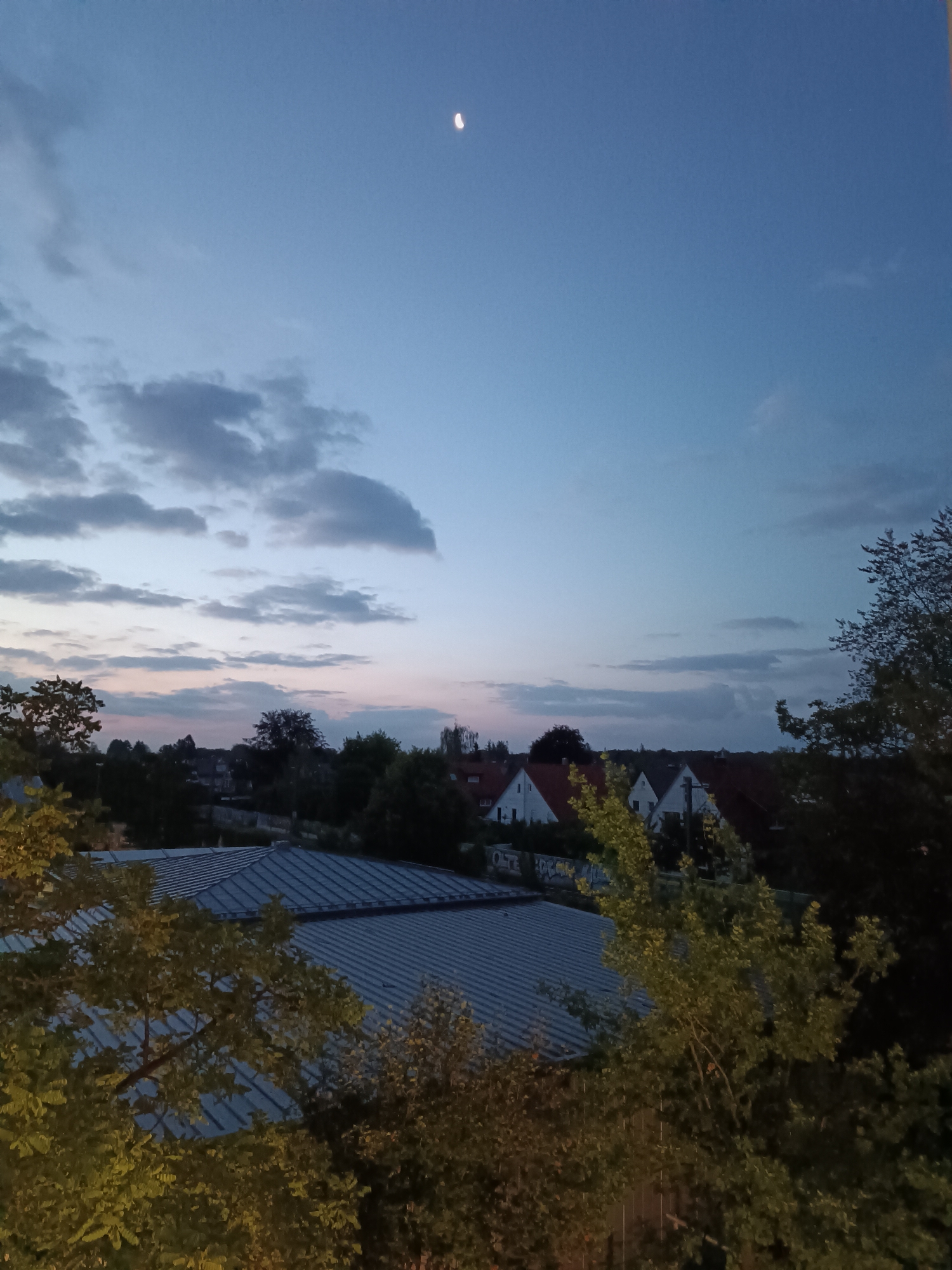 A twilight view of a suburban landscape in Germering, Germany, shows a clear sky with a crescent moon high above. Wispy clouds streak across the upper portion of the sky, transitioning to softer hues near the horizon where a faint pinkish glow suggests the last vestiges of sunset.
Below the sky, a collection of houses with red-tiled roofs are nestled amongst lush green trees. The buildings appear to be single-family homes, with several visible white facades. In the foreground, two large, leafy trees frame the scene, their branches extending towards the center. Behind the trees, a large, corrugated metal roof of what appears to be a commercial or industrial building dominates the lower part of the image. The overall impression is one of a peaceful evening in a residential area.