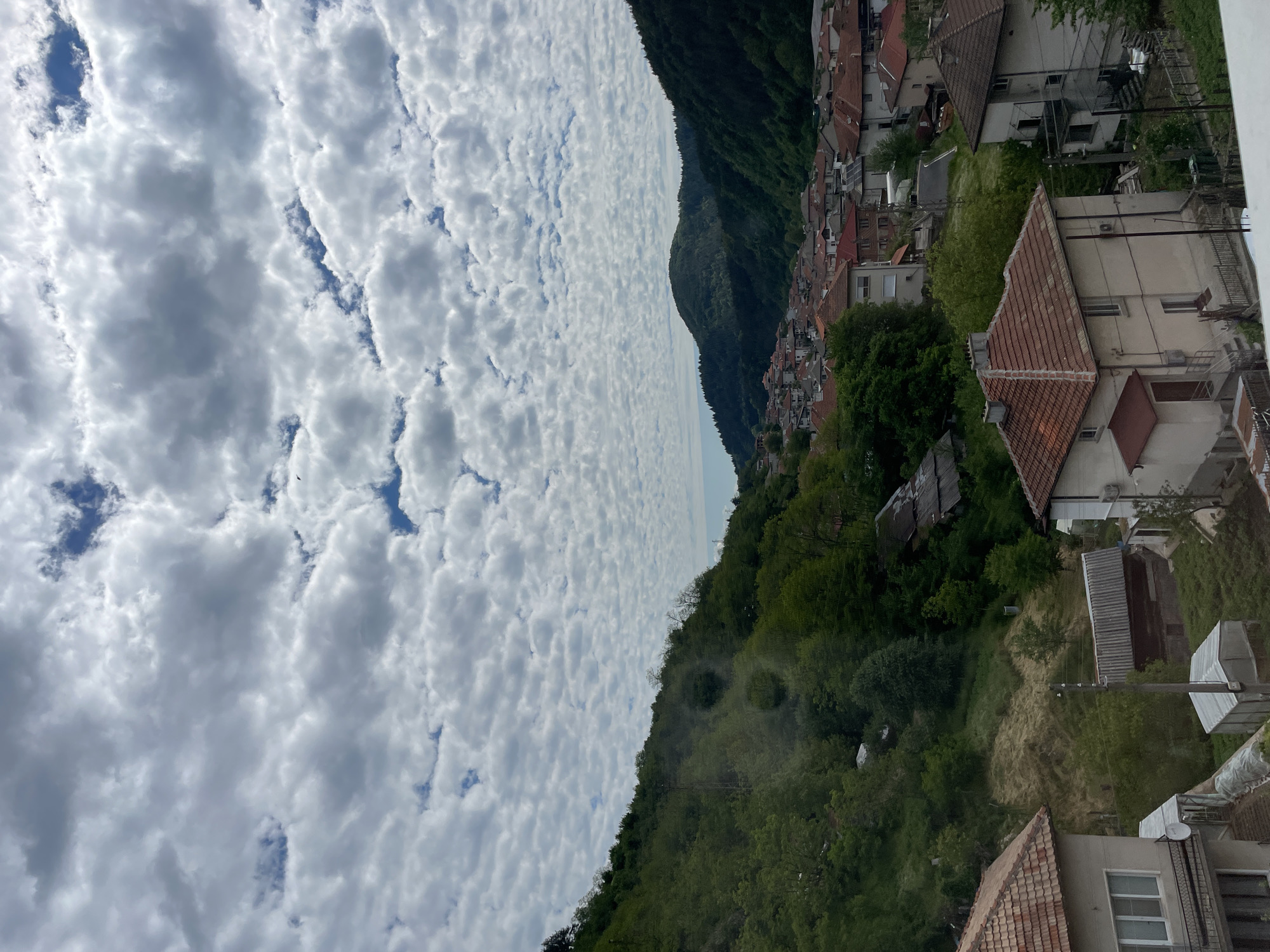 The image shows a view of Zlatograd, Bulgaria from an elevated position. The foreground consists of rooftops of buildings, which are predominantly covered with orange and red tiles. Beyond the buildings, there is dense green foliage, presumably trees and shrubs. A mountain range ascends into the distance, covered with more green vegetation. In the far distance, the landscape fades into the horizon with a faint line of blue suggesting the presence of water or sky meeting the land. The sky is filled with a field of altocumulus clouds, presenting a textured pattern of white and grey.