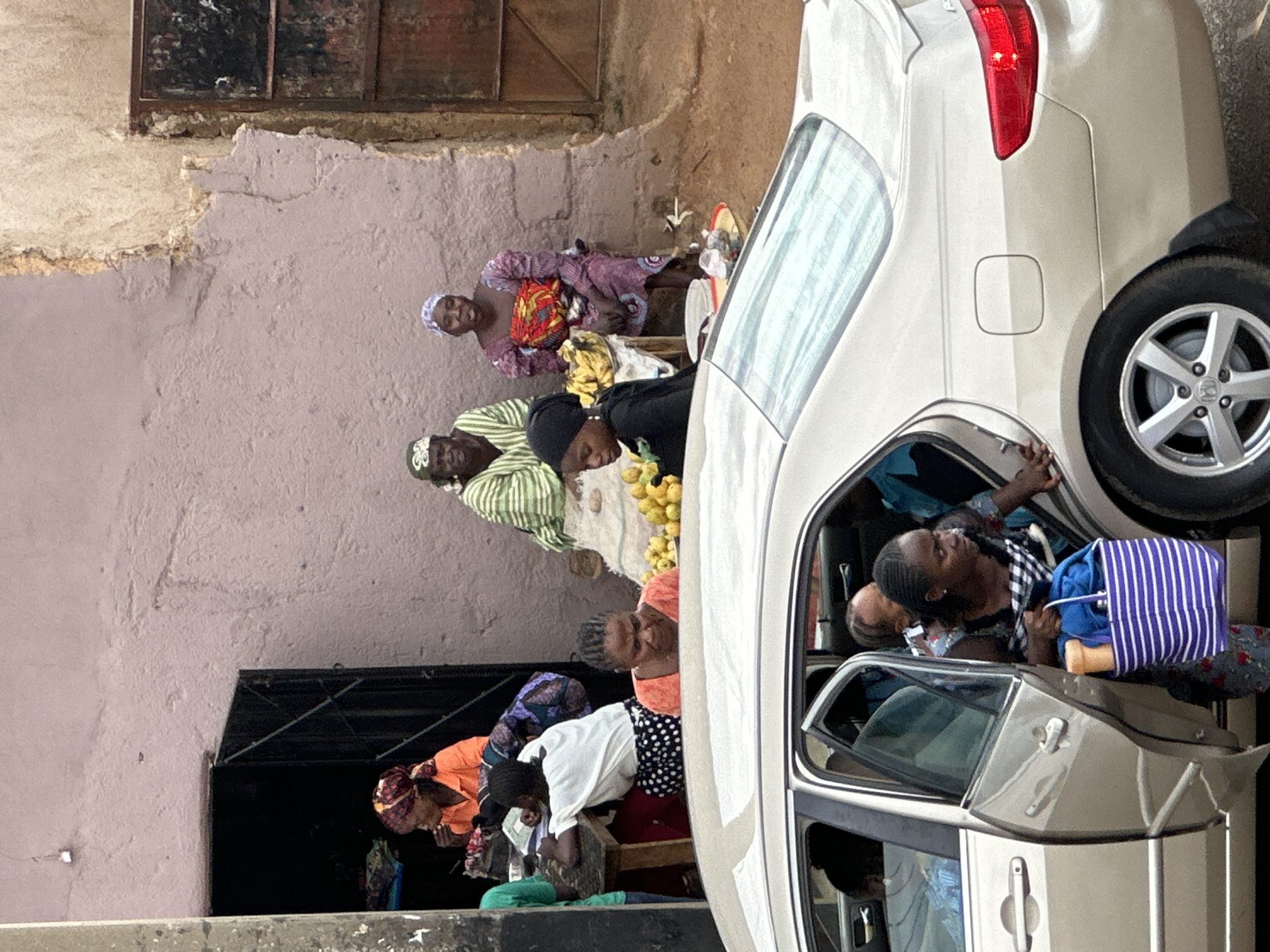 The image depicts a street scene in Jos, Nigeria, focused on a roadside market and a car. In the foreground, a light-colored sedan is parked with its passenger-side front door open. A woman, wearing a checkered top and carrying a striped purple and white tote bag filled with items, is exiting the car. Several other individuals are visible inside the car, some partially obscured.
Behind the car, a group of women are gathered around a table laden with produce, primarily yellow fruits. One woman in an orange top and headscarf is seen at the far left, seemingly tending to goods. Two other women sit behind the table; one wears a green and white striped outfit, and the other is in a purple patterned dress. Another woman, wearing a black headscarf, is seated at the table, looking down. The background shows a textured, pale pink wall with sections of exposed brick and a dark doorway with a metal gate. The overall impression is a busy, everyday scene at a local market.