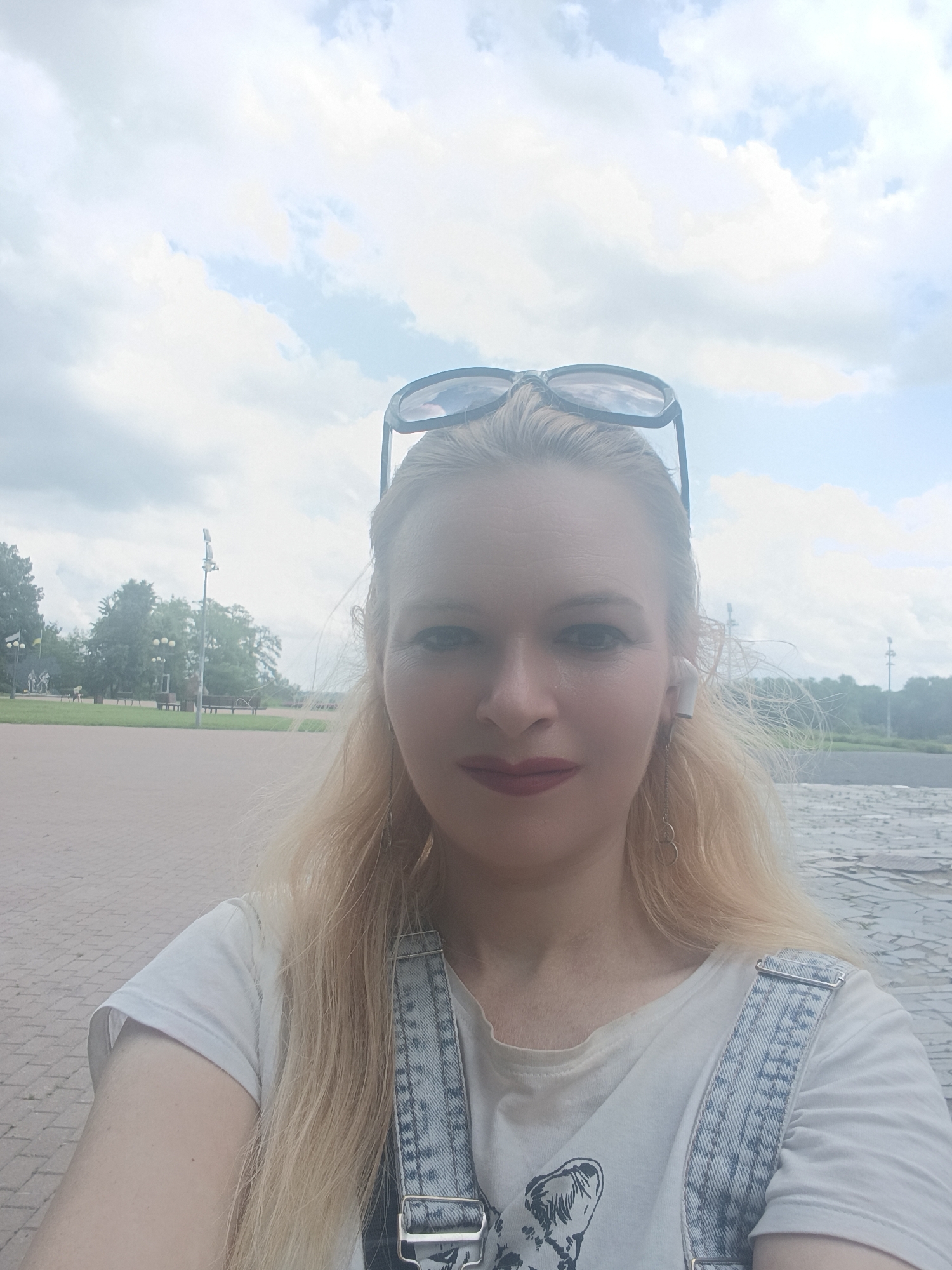 The image is a selfie taken outdoors in Chernihiv, Ukraine, on a cloudy day. A woman with blonde hair, wearing denim overalls over a white t-shirt, is the primary subject. She has sunglasses perched on her head and is wearing white wireless earbuds with a dangling earring. The background shows a paved area, trees, benches, and lamp posts, suggesting a park or public square. The overall lighting is soft due to the overcast sky.