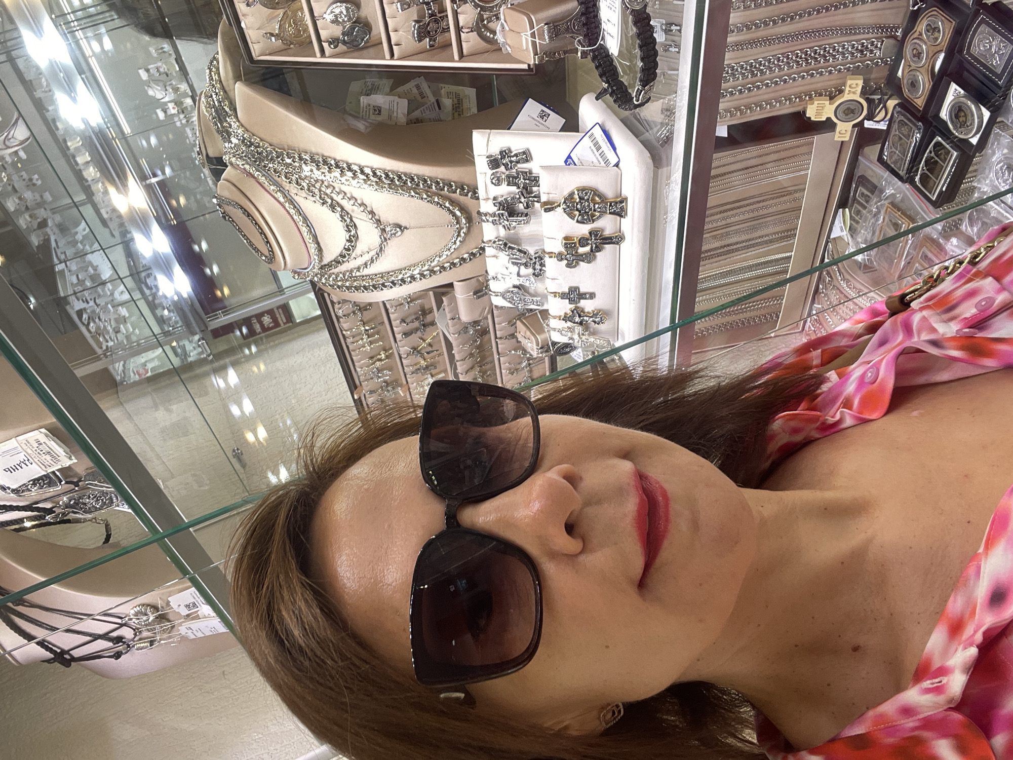 A middle-aged woman with brown hair, wearing large dark sunglasses, red lipstick, and a pink and white patterned shirt, takes a selfie in a jewelry store. She is positioned in the foreground, with various displays of silver necklaces and pendants visible behind her. The store appears well-lit, with glass display cases showcasing a wide assortment of jewelry, including numerous cross-shaped pendants and chains. The background reveals a reflection of herself and the store's interior, suggesting a mirrored surface or glass. The presence of retail displays and the nature of the merchandise indicate this is a commercial establishment, likely a jewelry shop. The context of Sochi, Russia, if this information is provided from an external source, implies the location of this retail setting.
