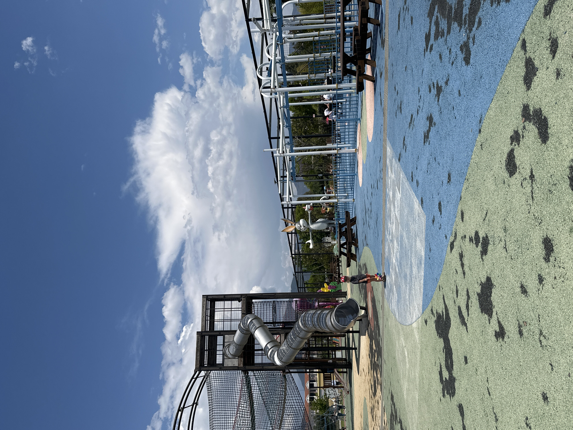 The image captures a vibrant outdoor playground in Fethiye, Turkey, set against a backdrop of a partly cloudy blue sky. The playground features colorful surfacing, including blue and green sections. Prominent playground structures include a metal climbing frame with a winding slide, and a metal play structure with overhead bars. A large Bugs Bunny statue stands on the playground.
A child is standing on the playground surface, looking toward the slide. In the background, other people can be seen on the higher metal play structure.