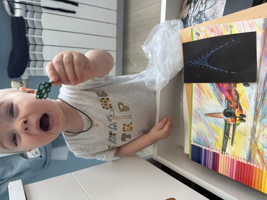 A frontal, waist-up view captures a young child within an indoor setting in Votkinsk, Russia. The child, with light-colored hair, wears a grey short-sleeved t-shirt featuring a 'SAVANNA TRIP' graphic and a black cord necklace with a small gold-colored cross. The child's mouth is wide open, mid-vocalization, with an expression of intense engagement, looking directly towards the camera. The child's left hand extends forward, holding a small, dark green, intricately shaped object resembling a decorative key or a multi-lobed form. The child's torso is positioned above an open white drawer, revealing its contents: a spiral-bound notebook with a colorful airplane illustration on its cover, a section displaying numerous colored pencil tips along its left edge, and a smaller black rectangular piece of material adorned with a blue string art design resembling an arched structure on top. A crumpled transparent plastic bag is also visible within the drawer. In the background, a white radiator is affixed to a light blue wall. The floor appears to be a light wood or laminate.