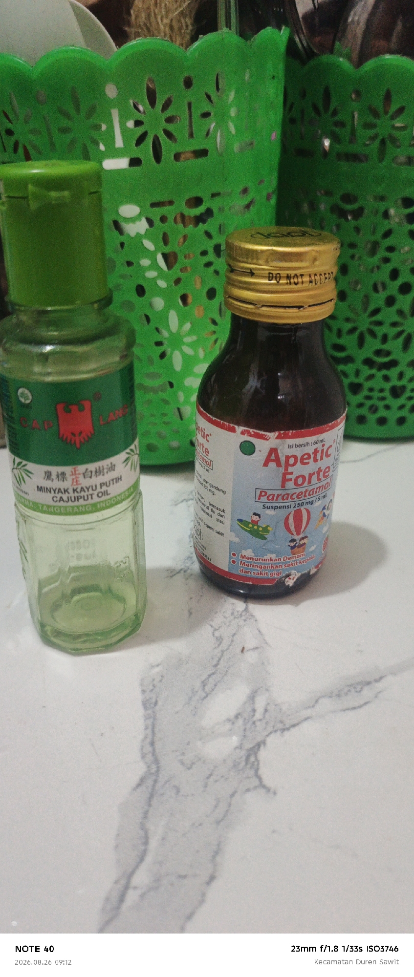 The image shows two bottles of medication on a white marble-patterned surface. The bottle on the left is a small green glass bottle with a green cap, labeled "CAP LANG MINYAK KAYU PUTIH CAJUPUT OIL". The bottle on the right is a dark glass bottle with a gold cap, labeled "Apectic Forte Paracetamol". Both bottles appear to be medicinal. In the background, there are two green plastic baskets. The bottom left of the image contains text that reads "NOTE 40 2026.08.26 09:12". The bottom right of the image contains text that reads "23mm f/1.8 1/33s ISO3746 Kecamatan Duren Sawit". The setting appears to be indoors, likely a home or pharmacy, given the products and baskets. The text "Kecamatan Duren Sawit" indicates the location as Duren Sawit Subdistrict, East Jakarta, Indonesia.