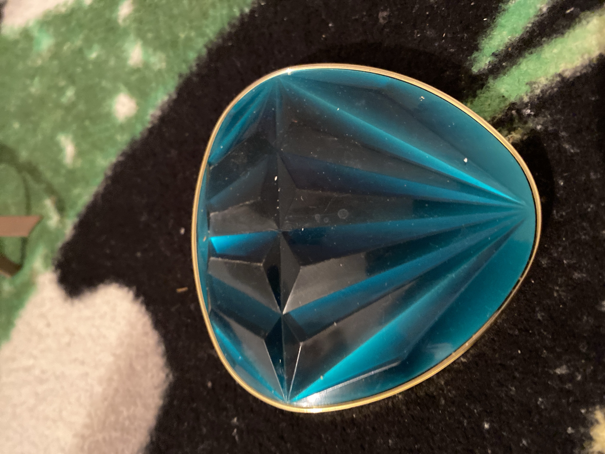This is a close-up, overhead shot of a faceted, teal-colored object with a gold rim. The object is oval and appears to be made of glass or plastic. Its surface is cut with numerous facets, creating a pattern that resembles a gemstone or a stylized fan. The object is lying on a dark, textured surface that has some lighter, fuzzy patches and faint green streaks. The background is dark and blurry, with some lighter speckles. There is no text visible in the image. The lighting suggests an indoor setting, possibly artificial, as there are some highlights reflecting off the object's surface. The image is focused on the object, with the background out of focus.