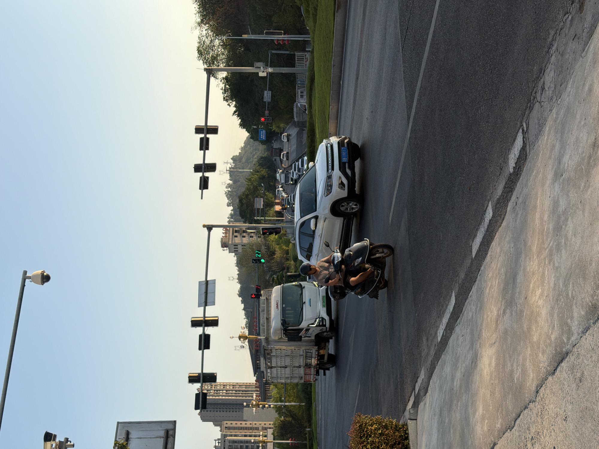 In Jiangjin, China, at an intersection during daylight, a scooter rider wearing a helmet and a brown t-shirt navigates through traffic. They are positioned on the right side of the image, mid-turn. To their left, a white truck with a cargo bed is visible, with its license plate reading "B 04999". Further in the background, a white SUV is seen with the license plate "B 19Y17". Traffic lights are present above the road, with some displaying red and green signals. The environment includes asphalt roads, a concrete sidewalk on the left with a small bush, and buildings and trees in the distance under a clear sky.