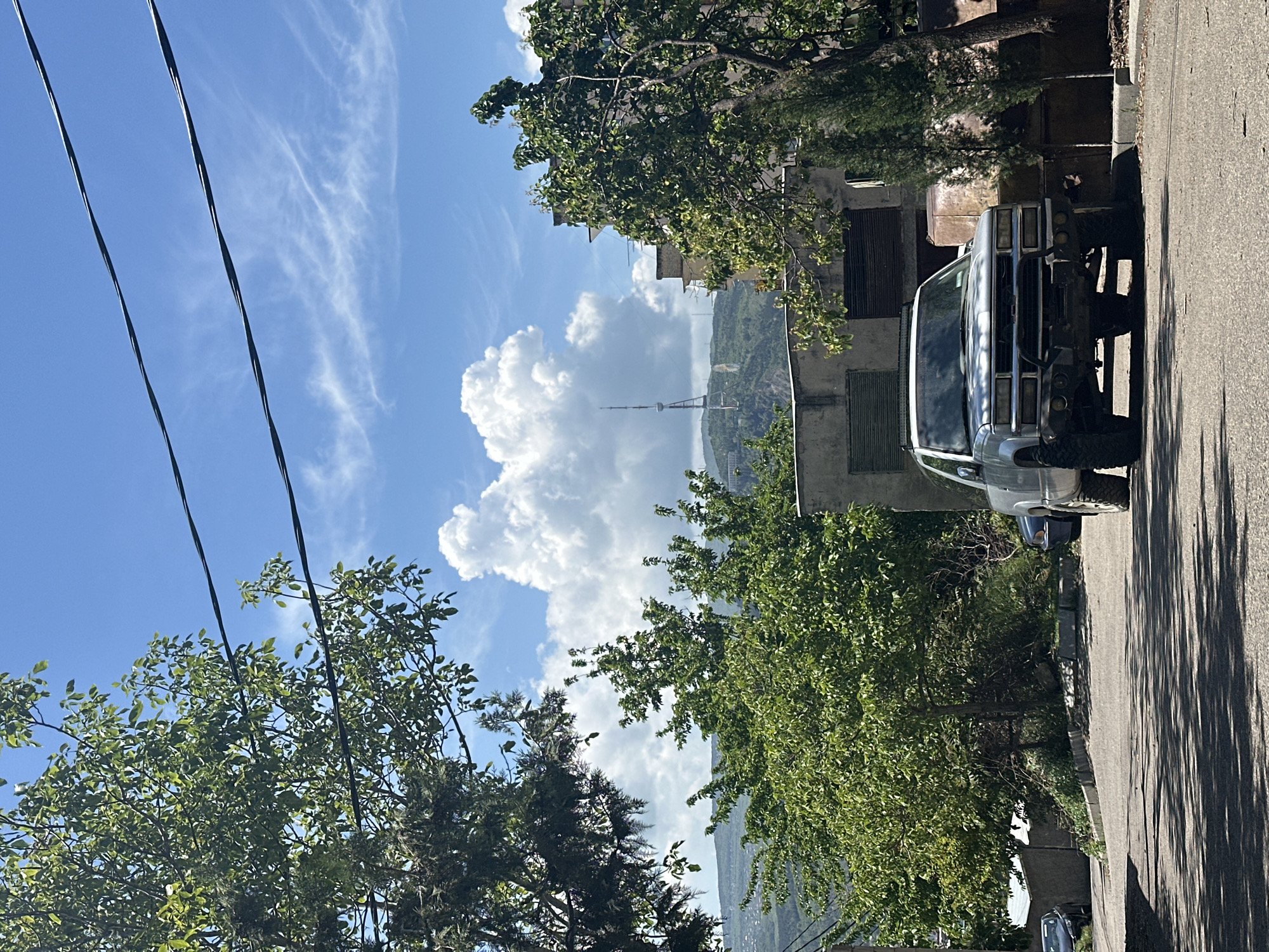 The image shows a street scene in Tbilisi, Georgia, with a view of the Tbilisi TV Broadcasting Tower in the distance. A silver SUV is parked on the side of the road. Trees line the street, partially obscuring the view of the tower and the sky. Power lines are visible in the upper left corner of the frame. Buildings are visible behind the trees and the car. The sky is blue with white clouds. The image is taken at an angle, appearing to be shot from a slightly elevated position.