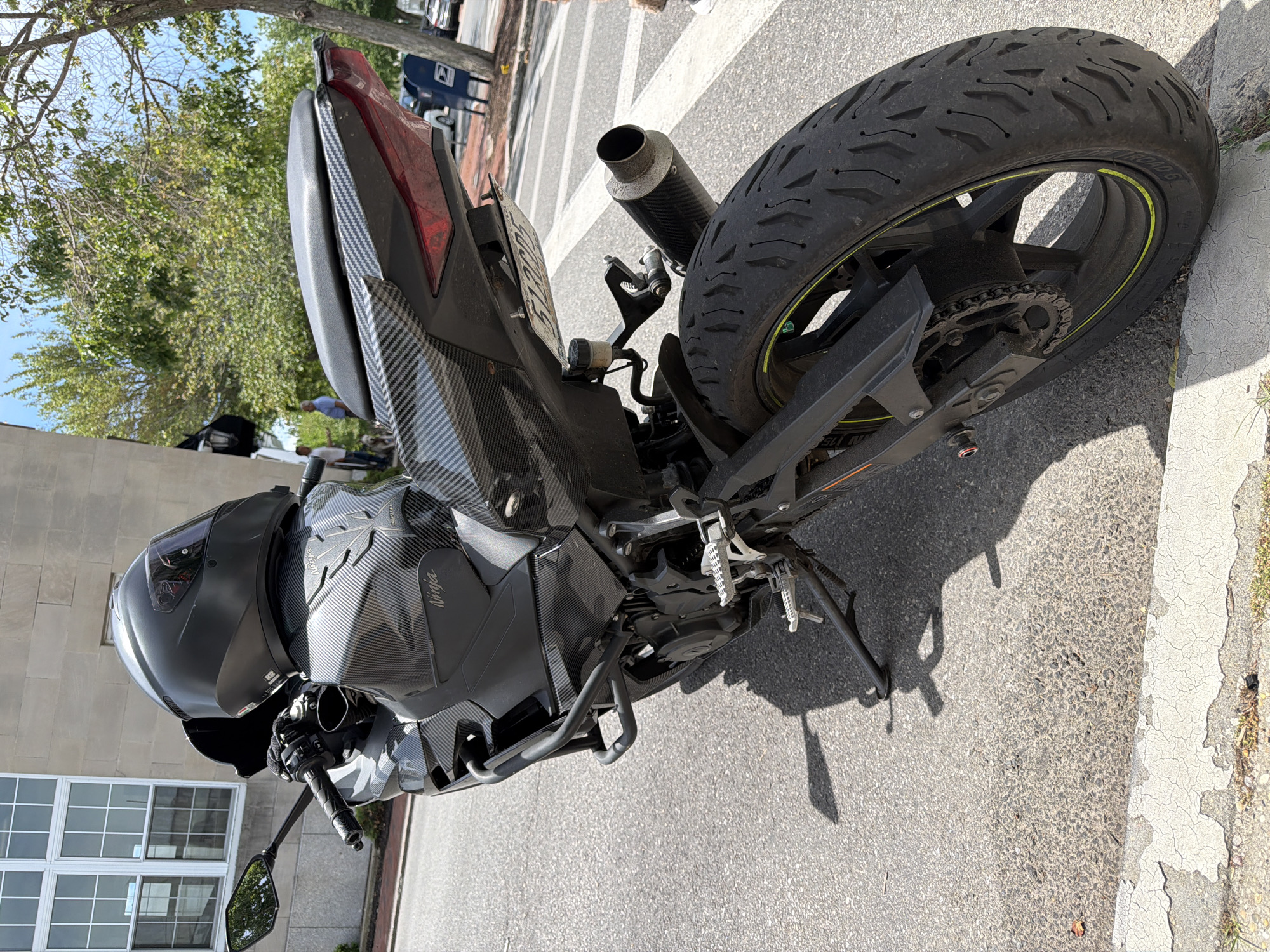 A black Kawasaki Ninja motorcycle is parked on a street in the Village of Southampton, United States of America. The motorcycle is adorned with carbon fiber accents on the fuel tank, tail section, and fenders. A black helmet rests on the seat, and a license plate is attached to the rear. The motorcycle is situated on an asphalt surface with a cracked white painted curb and sidewalk. In the background, a brick building with large windows and lush green trees are visible. Two men and a dog are standing on the sidewalk in the distance, and a blue mailbox is also present. The scene is illuminated by bright sunlight, casting shadows on the ground.