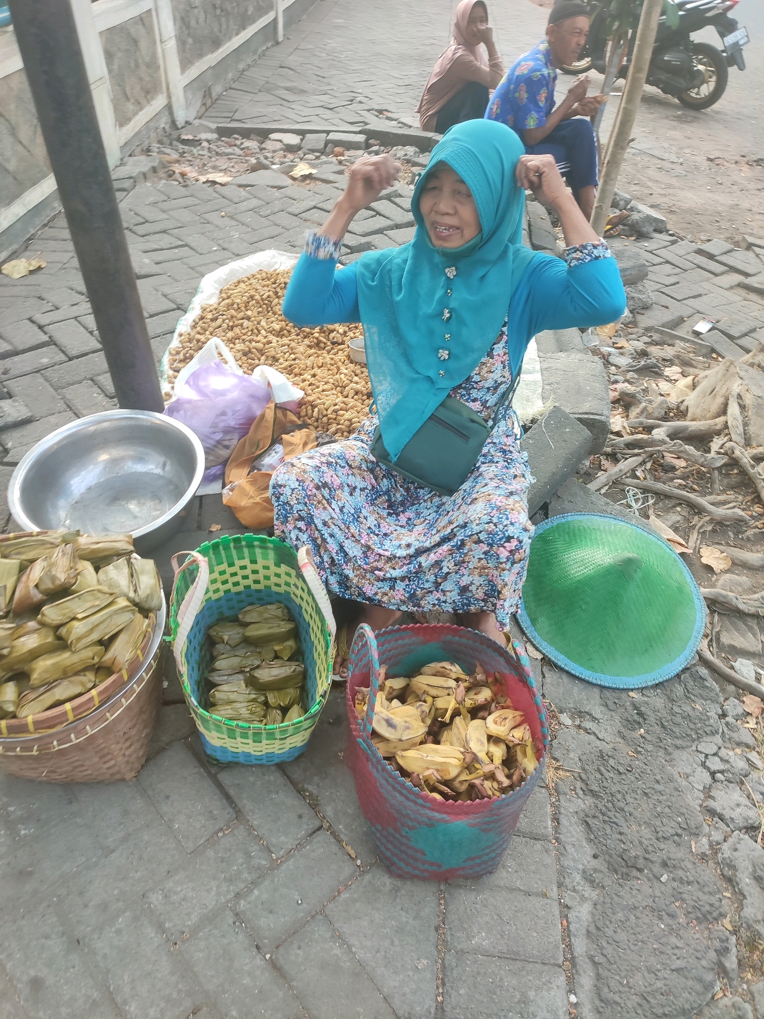In Nganjuk, Indonesia, a woman in a turquoise hijab and a floral dress sits on a paved sidewalk next to a large sack of peanuts. She is actively gesturing with her hands, with a visible smile on her face. In front of her are several woven baskets filled with food items wrapped in banana leaves. To her left, a large metal bowl sits empty. A green conical hat rests on the ground to her right. In the background, two individuals, one wearing a blue shirt and the other a light brown hijab, are seated. A motorcycle is parked nearby. The environment appears to be an outdoor market or street vendor setting.