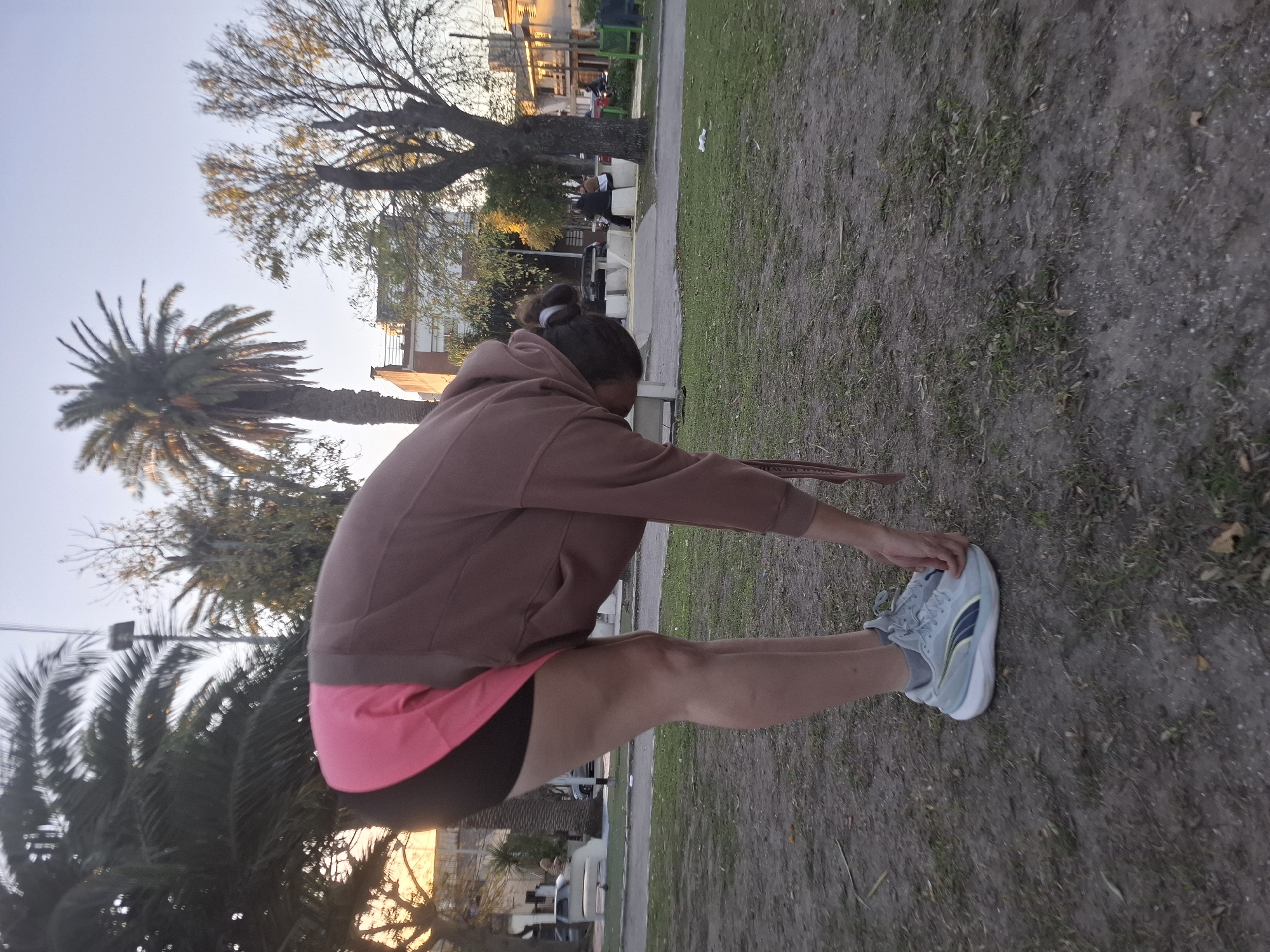 A person is captured mid-stretch in an outdoor park setting, likely in Villa Domínico, Argentina, during the late afternoon or early evening. The individual, seen from a slightly elevated angle, is bending forward at the waist, reaching towards their feet or shins, indicating a warm-up or cool-down exercise. They are wearing a brown hooded sweatshirt, pink shorts layered over black shorts, and light blue/grey sneakers with a dark blue and yellow stripe detail. Their hair is tied back in a bun.
The background reveals a public green space with a mix of earthy ground, sparse grass, and a clear dirt path. Various trees are present, including tall palm trees and deciduous trees, some with autumn-like foliage. In the middle ground, there are white benches where other people can be vaguely seen sitting. Beyond the park, residential buildings with some architectural details are visible, bathed in the warm, soft light characteristic of dusk or dawn. The sky is bright but hazy, with no visible clouds. No visible text is present in the image.